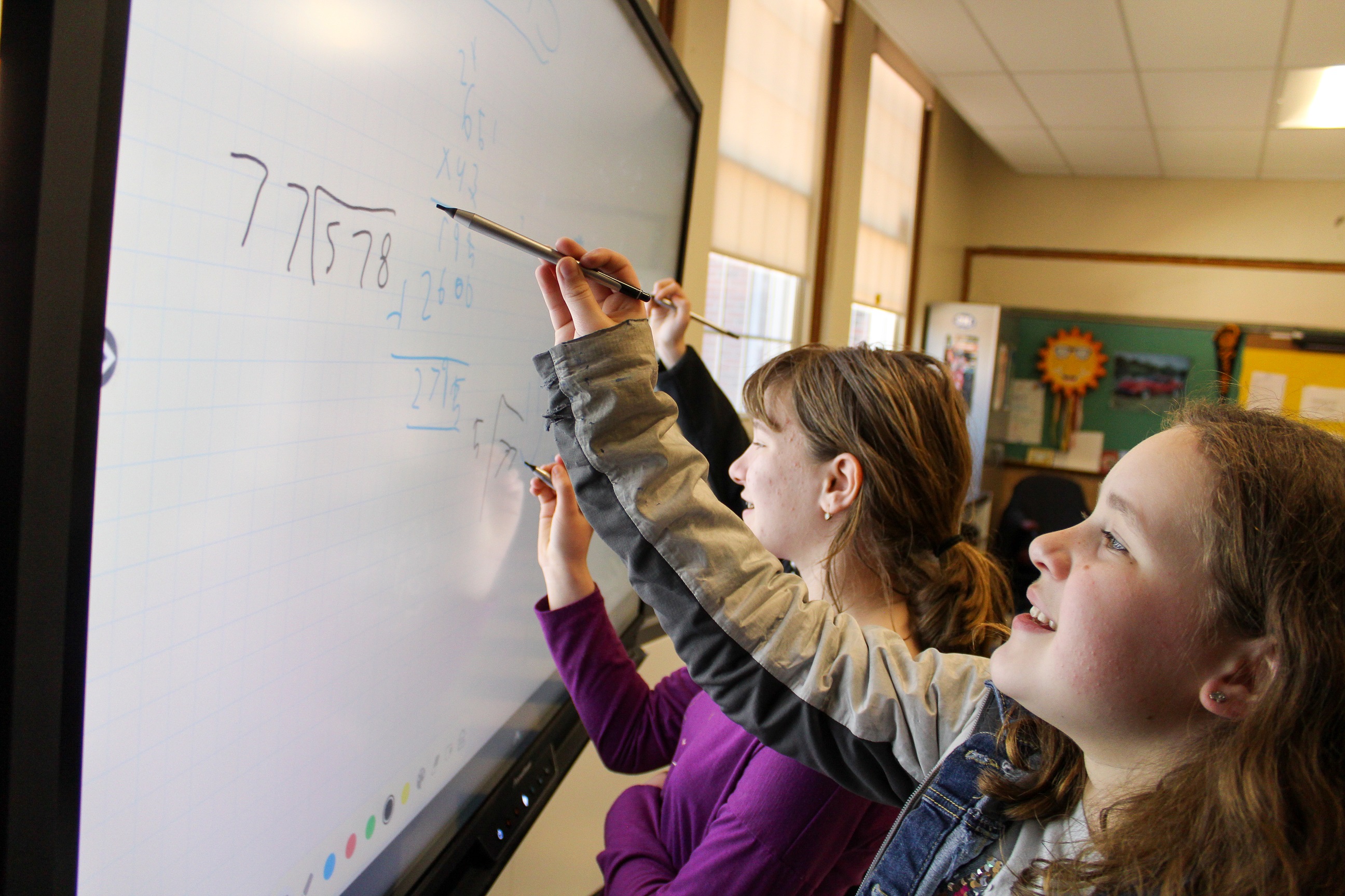two elementary school girls work on long division and multiplication using an interactive whiteboard