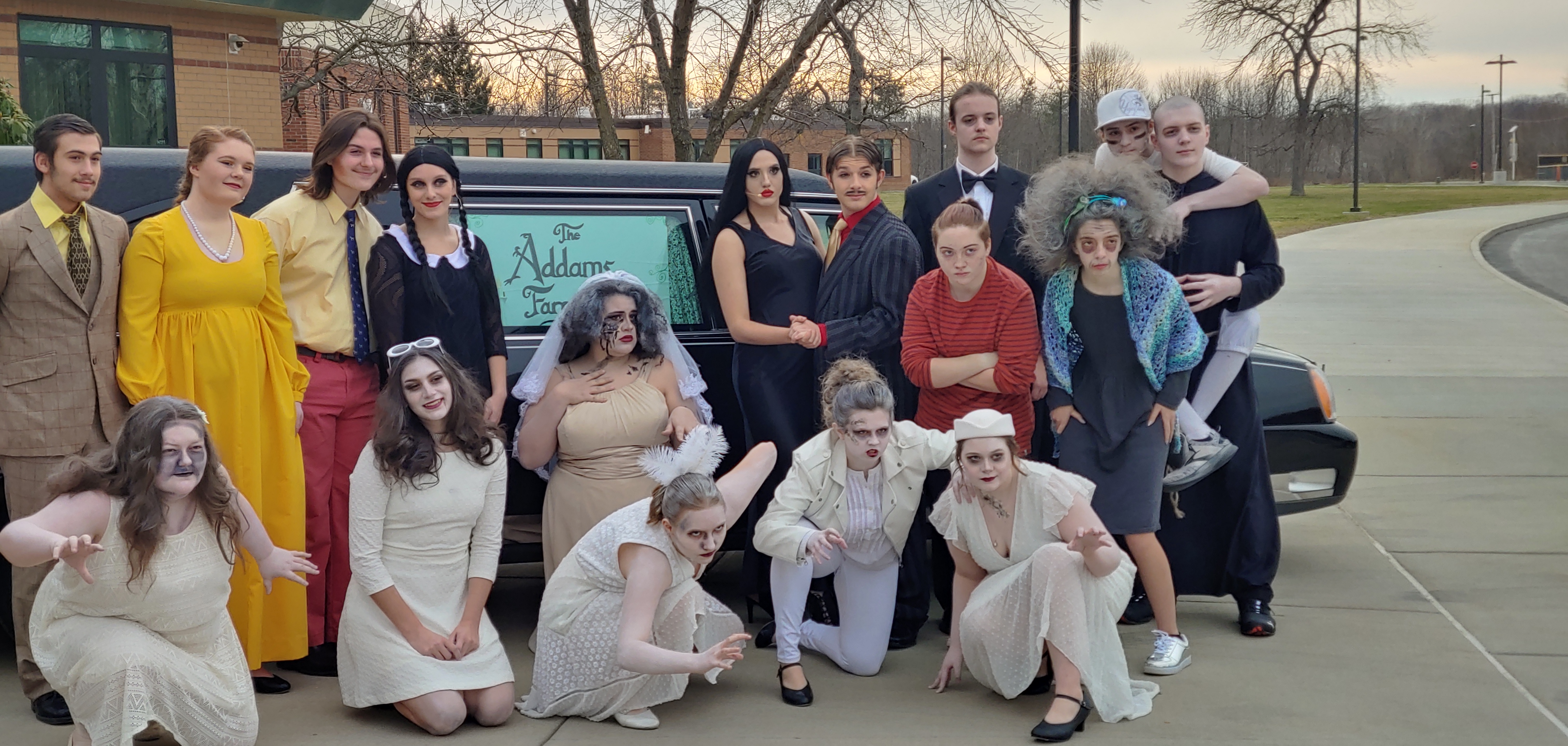 Cast of The Addams Family musical pose in costume in front of a hearse 