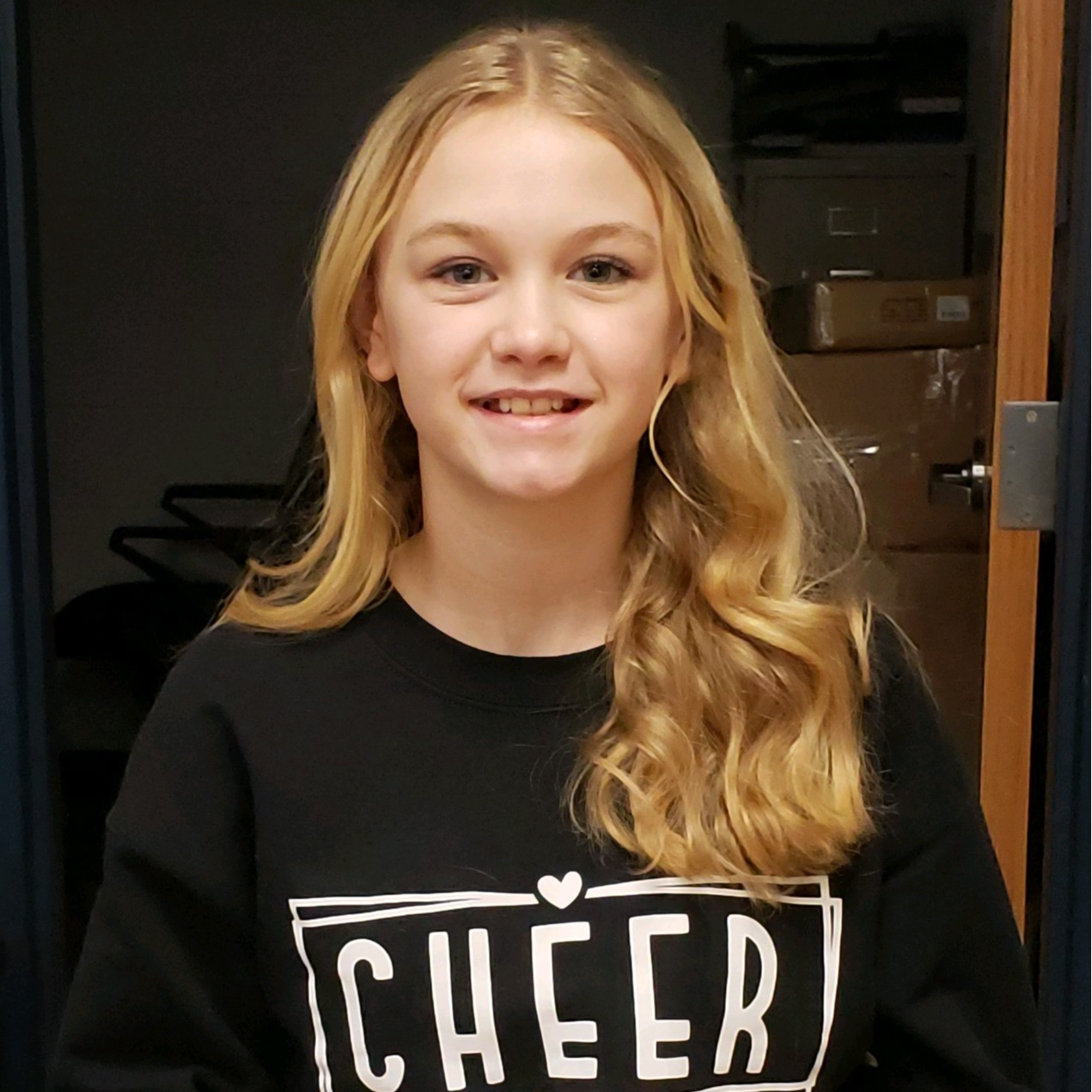 smiling girl with long blonde hair wearing a black sweatshirt with the word "CHEER""