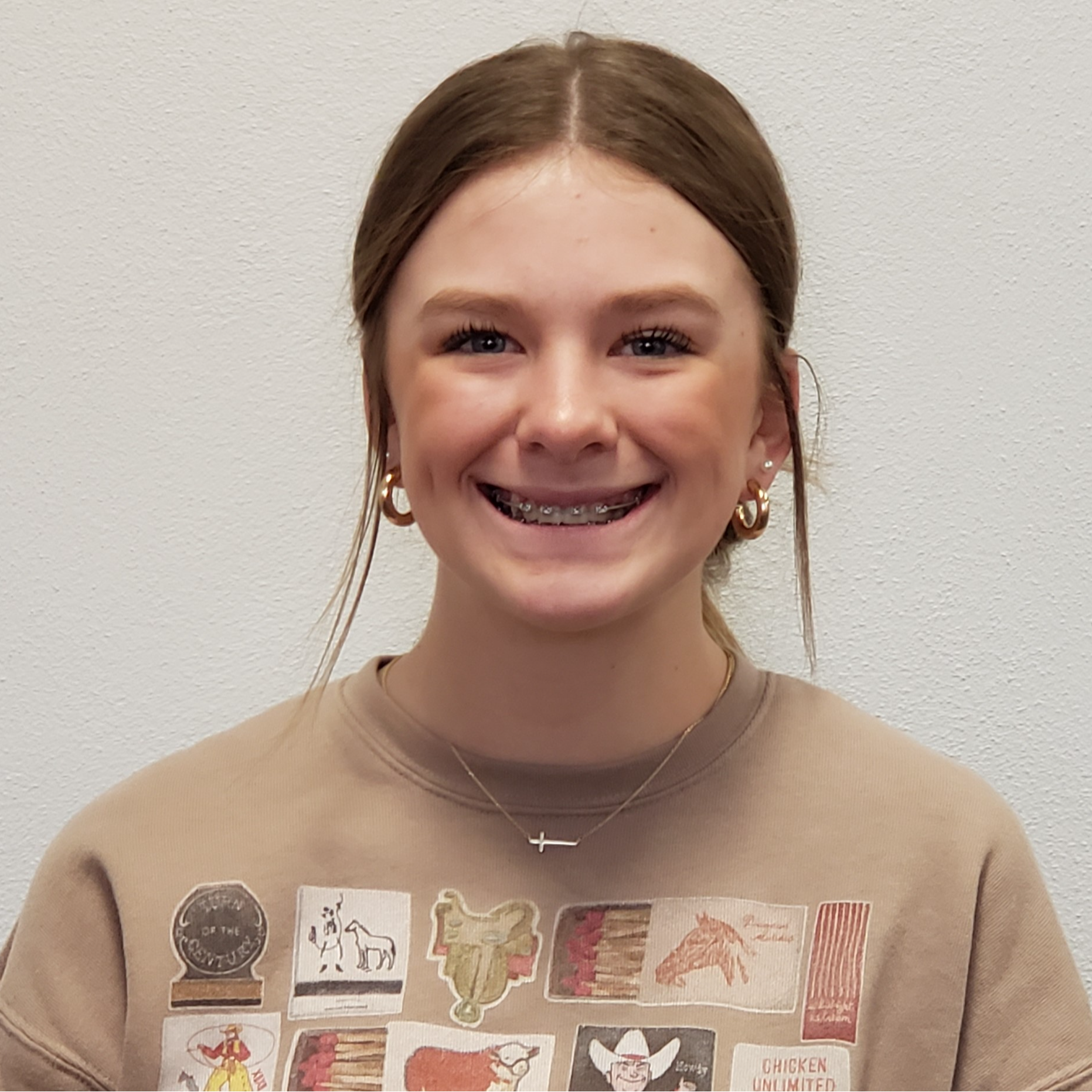 smiling girl with dark blonde hair pulled up wearing a tan sweatshirt with pictures of saddles, horses, cowboys, and cows on it