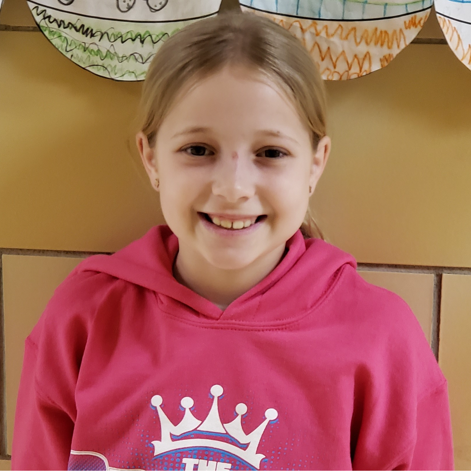 smiling girl with blonde hair pulled back wearing a pink hooded sweatshirt with a white crown on it