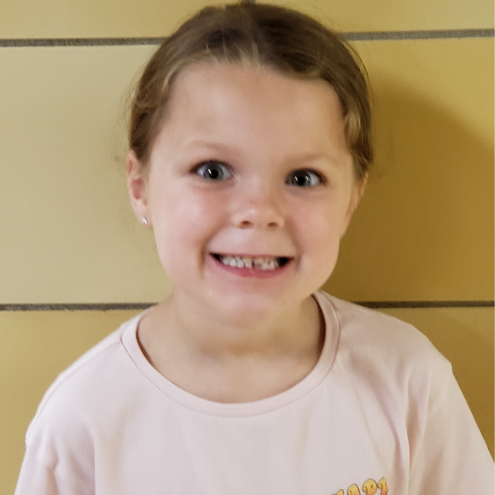 smiling young girl missing a front tooth with light brown hair pulled back wearing a light pink t-shirt