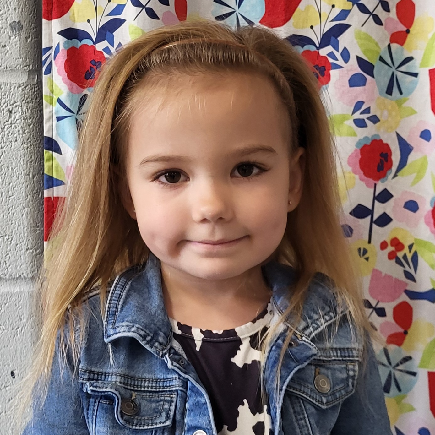 smiling little girl with dark blonde hair pulled back with a headband wearing a black & white splotched shirt under a jean jacket and standing in front of a flowered background