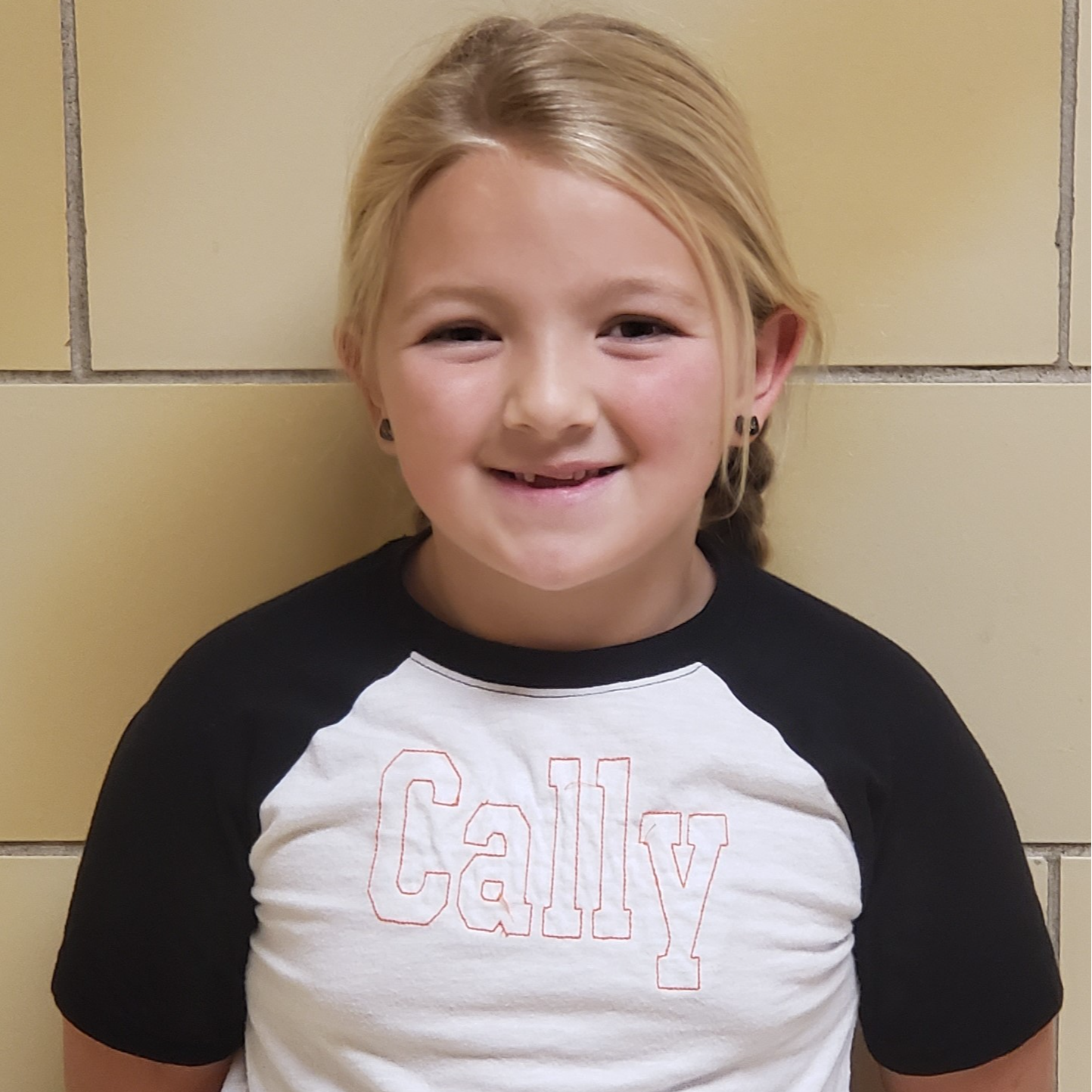 smiling girl with blonde hair  pulled back in braids wearing a white t-shirt with black sleeves and black earrings