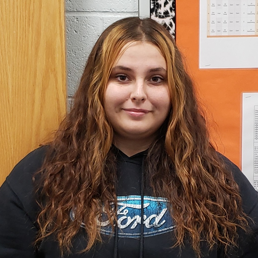 young lady with brown wavy hair with highlights in the front wearing a black hoodiege silver cross necklace, and glasses