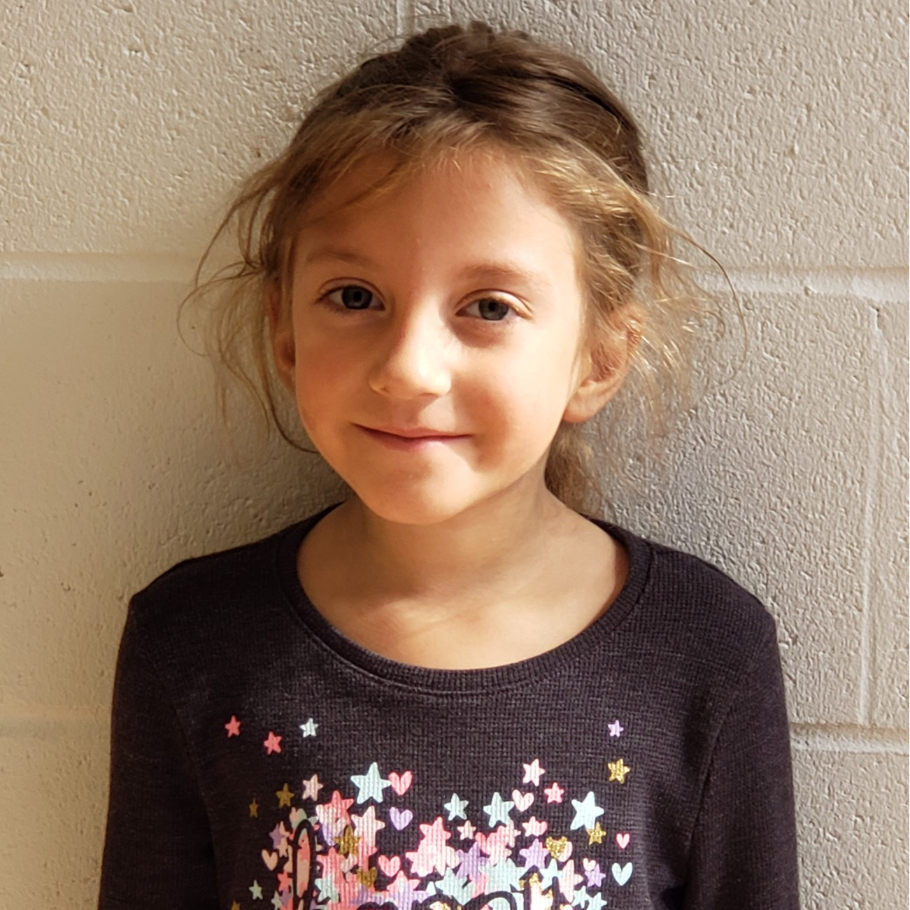 little girl with dark blonde hair pulled back wearing a dark gray sweater with pink and blue hearts and stars on it