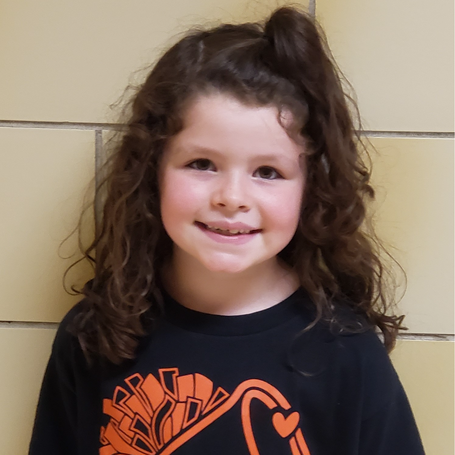 young girl long, curly brown hair with the top pulled up wearing a black t-shirt with orange poms on it