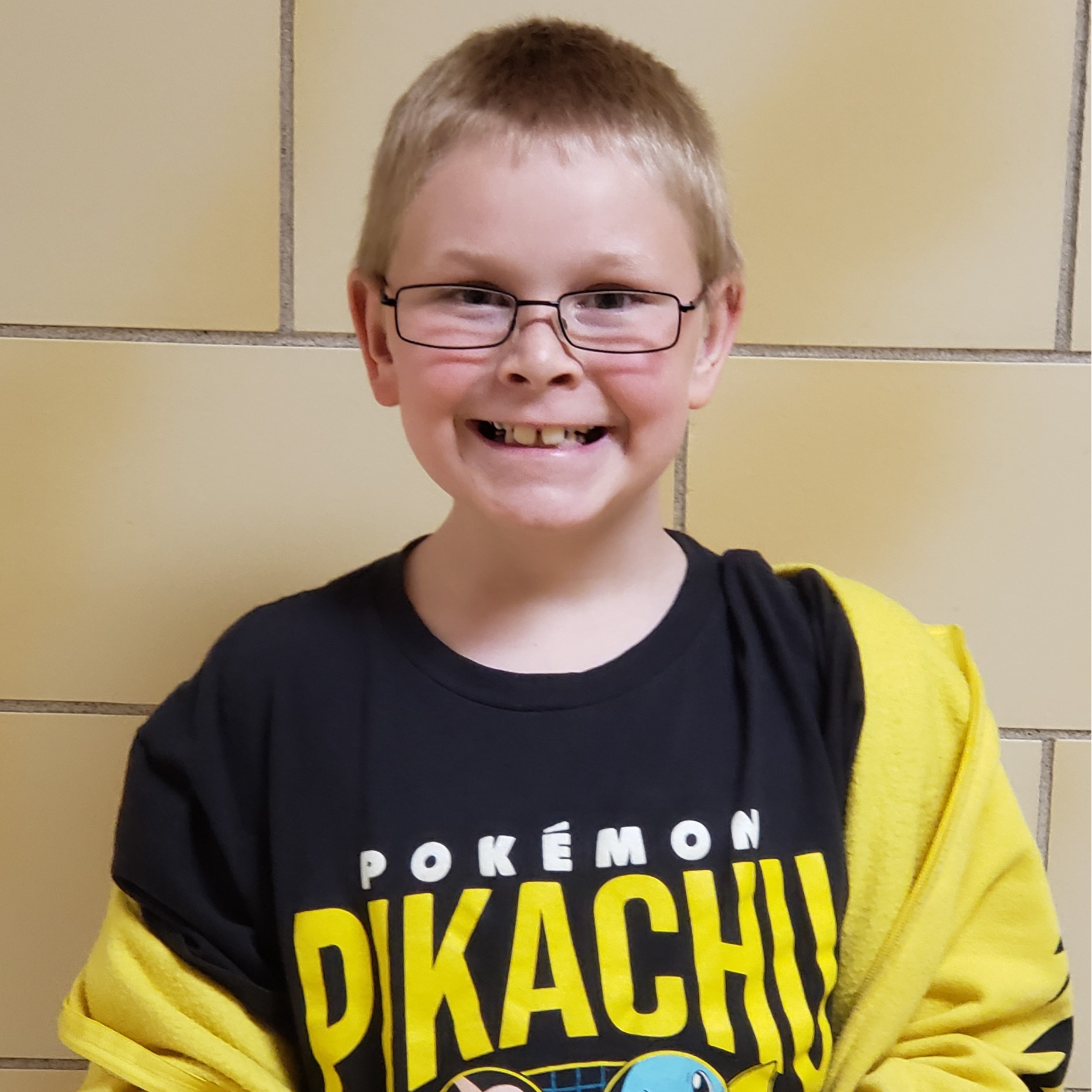 smiling boy with blonde hair and glasses wearing a black Pokemon Pikachu t-shirt under a yellow jacket that is falling off one shoulder