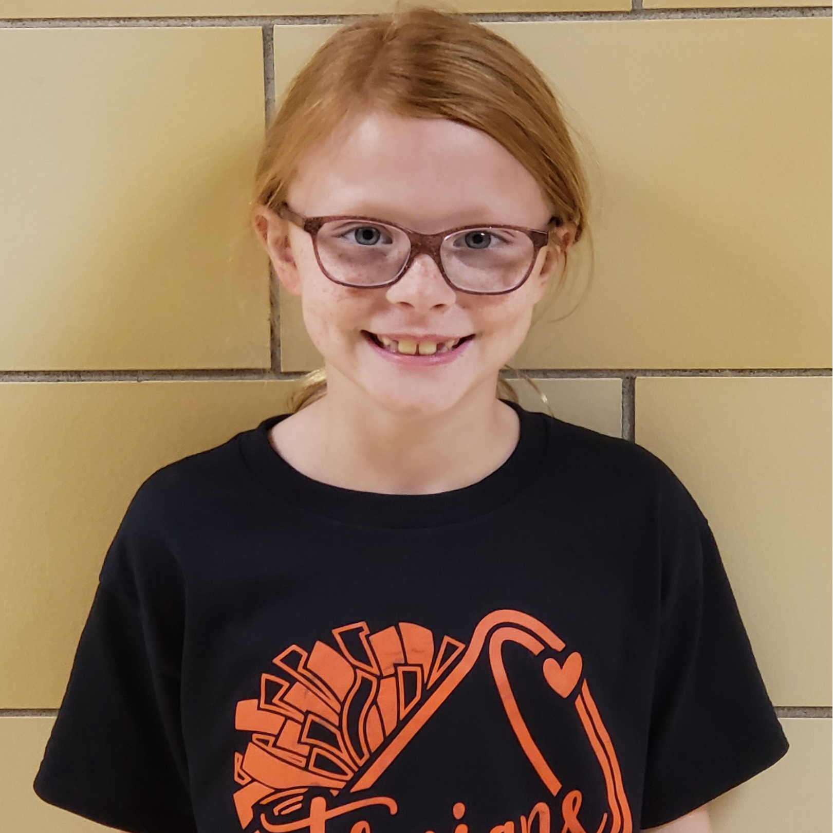 smiling girl with red hair pulled back  and glasses wearing a black t-shirt with orange poms on it