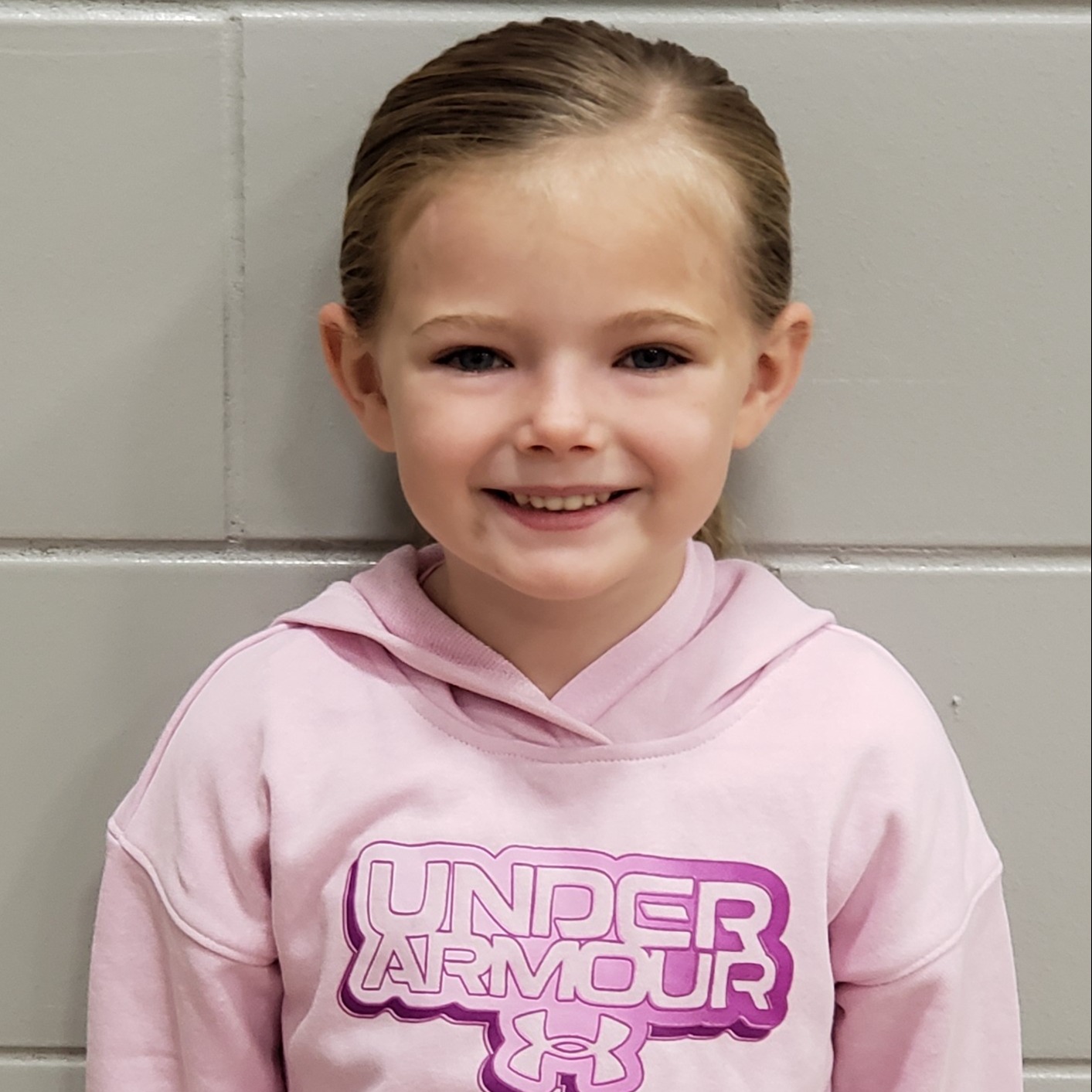 smiling little girl with dark blonde hair pulled back wearing a pink Under Armour Hoodie