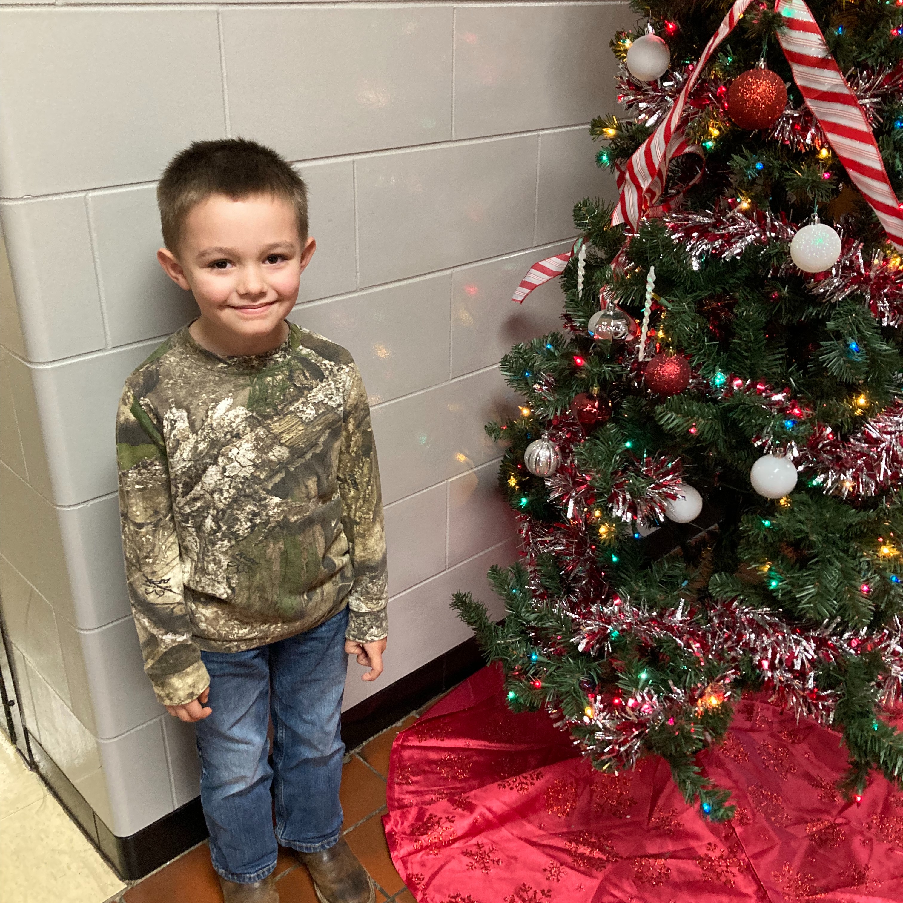 smiling young boy wearing a cammo shirt standing next to a christmas tree