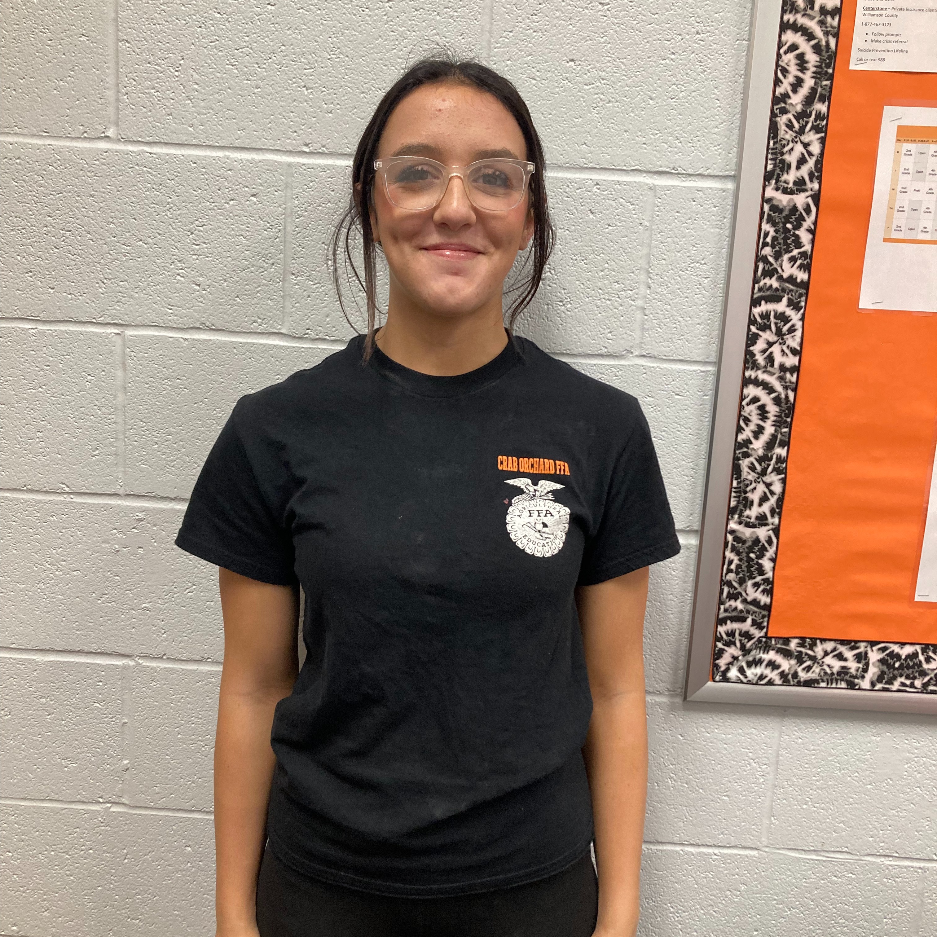 HS student with dark hear pulled back wearing clear glasses and a black, orange and white FFA t-shirt