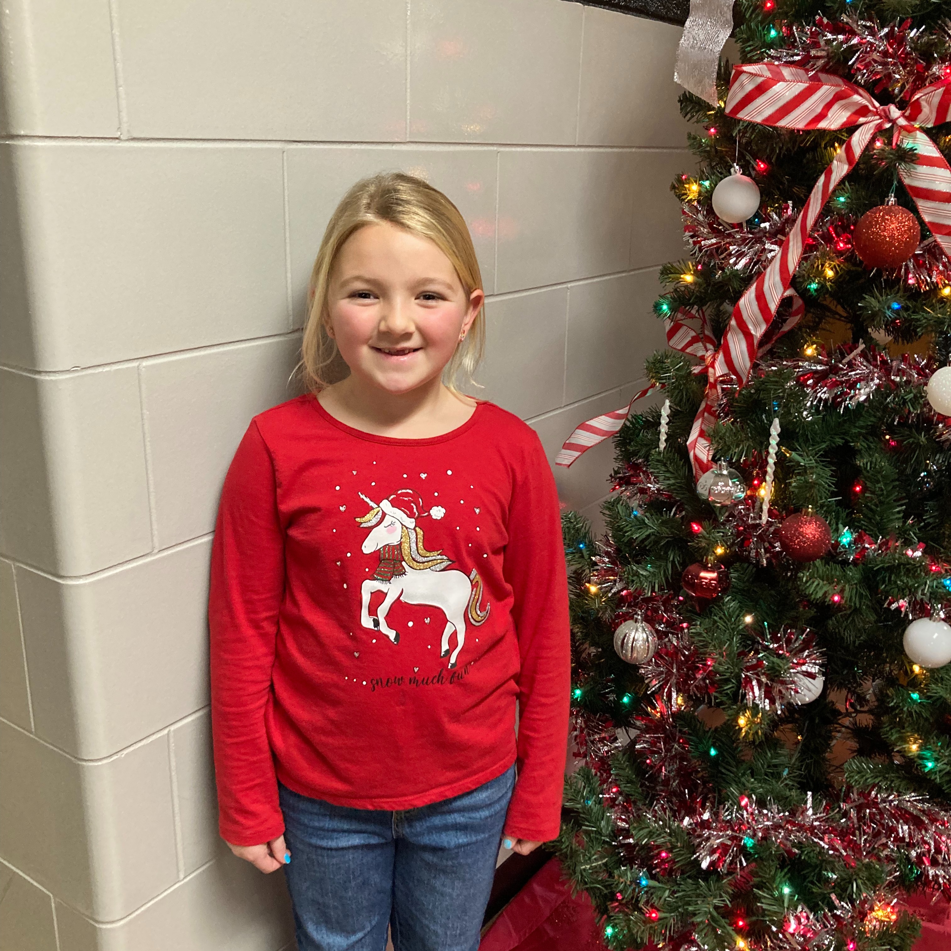 smiling little girl with blonde hair wearing a red and white unicorn christmas shirt standing next to a christmas tree