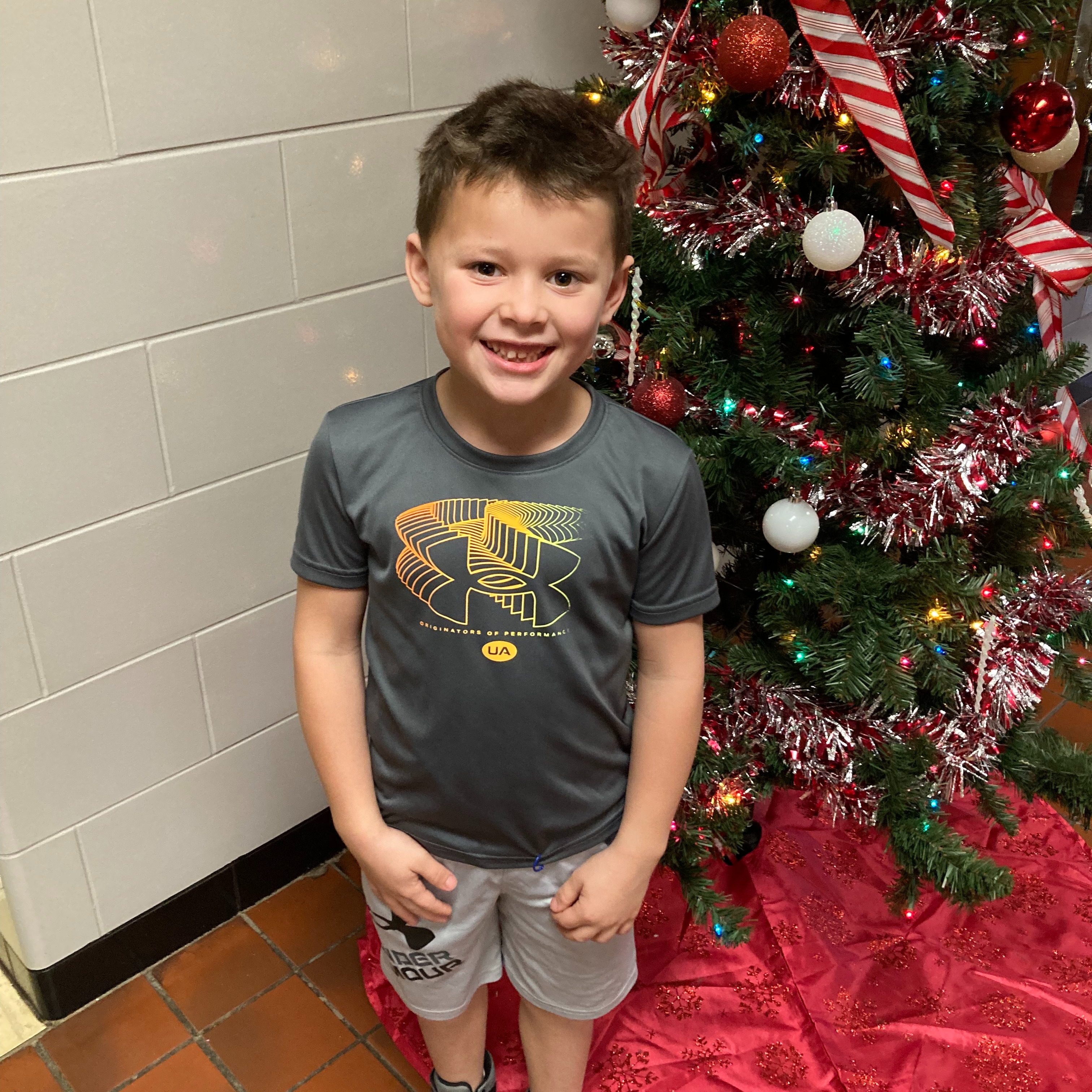 smiling little boy with dark hair wearing a grey orange and yellow under armour t-shirt standing next to a christmas tree