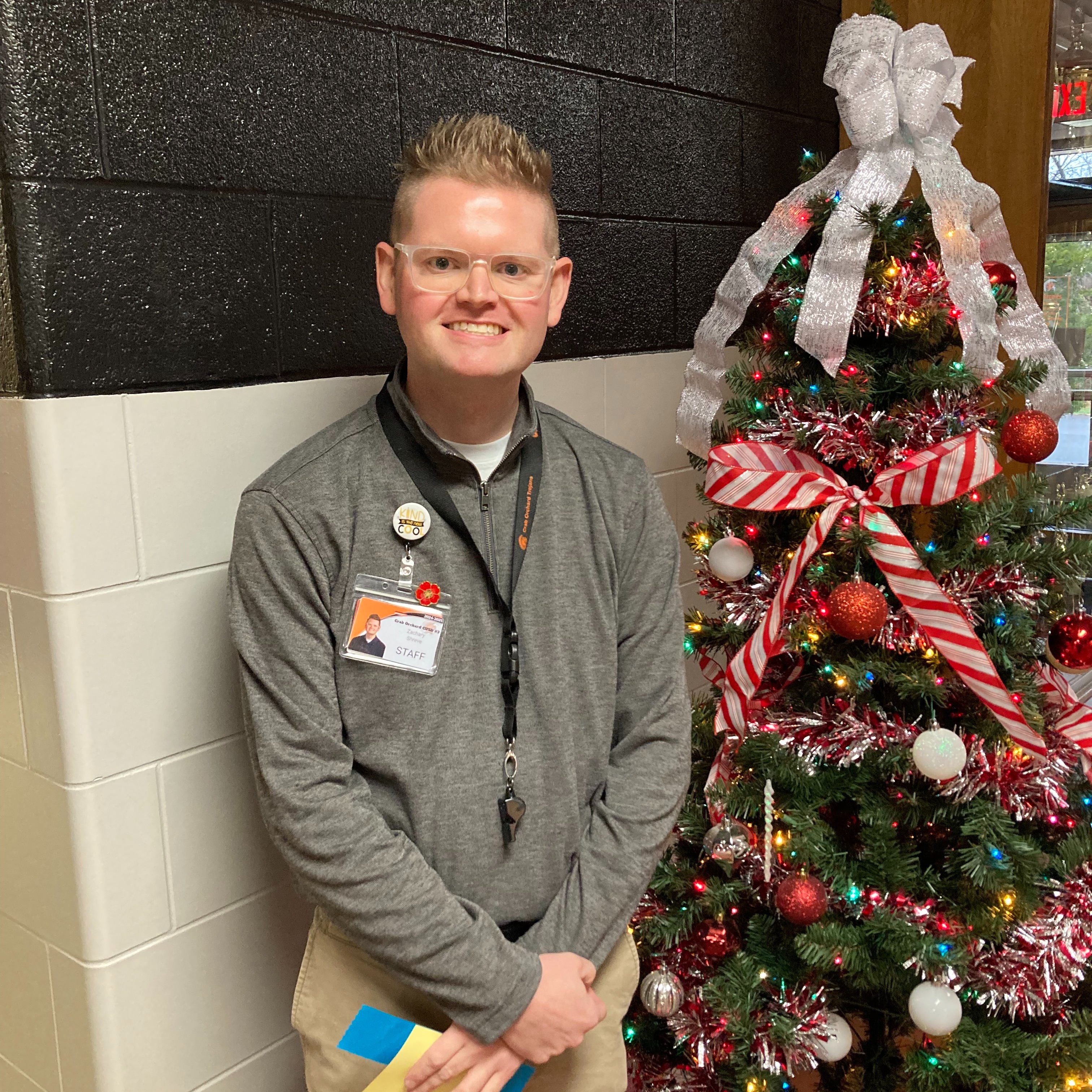 smiling male staff member wearing clear glasses, a grey long sleeve quarter zip shirt, a staff badge , and a black lanyard