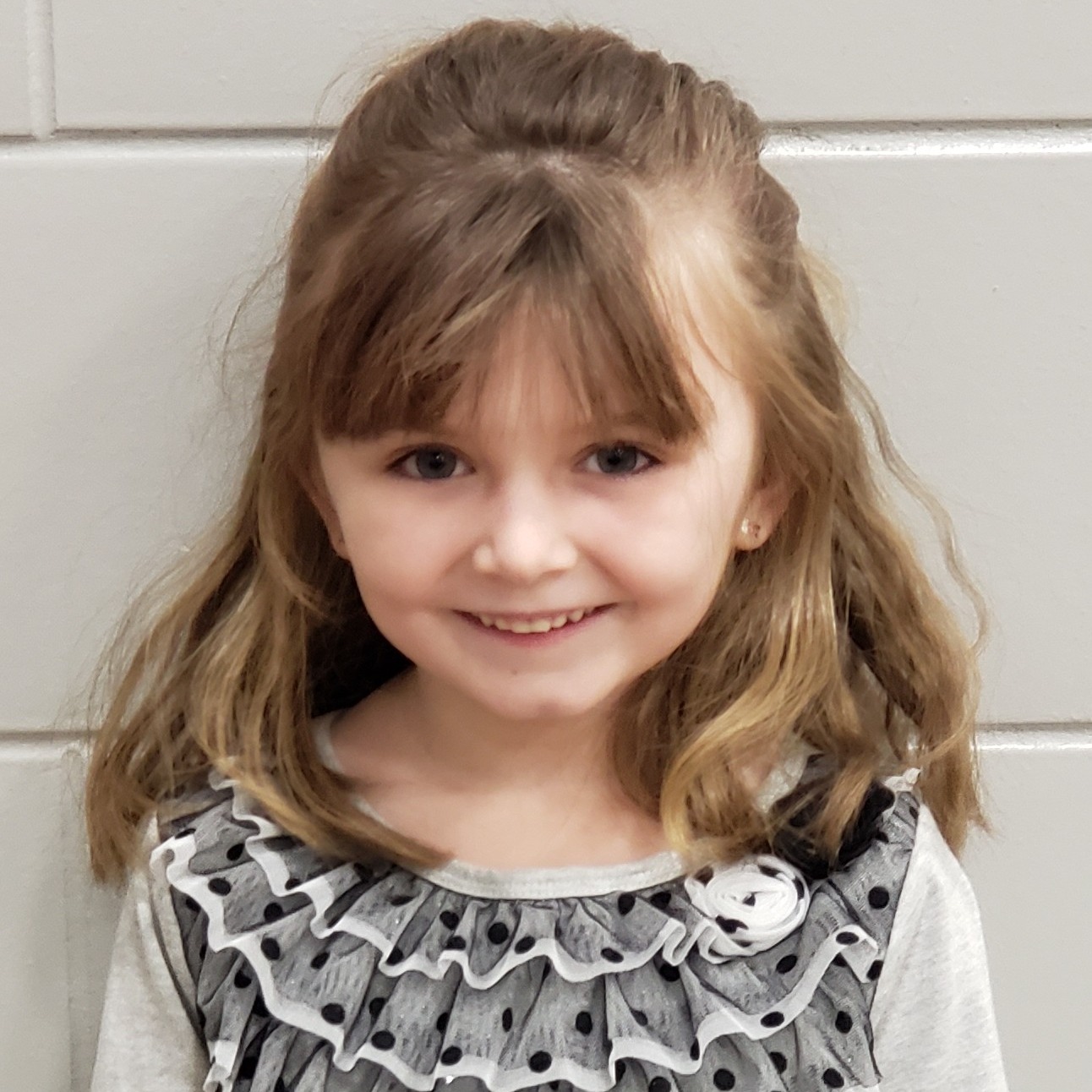 smiling young girl with dark blonde hair wearing a gray dress with ruffles and black polka dots