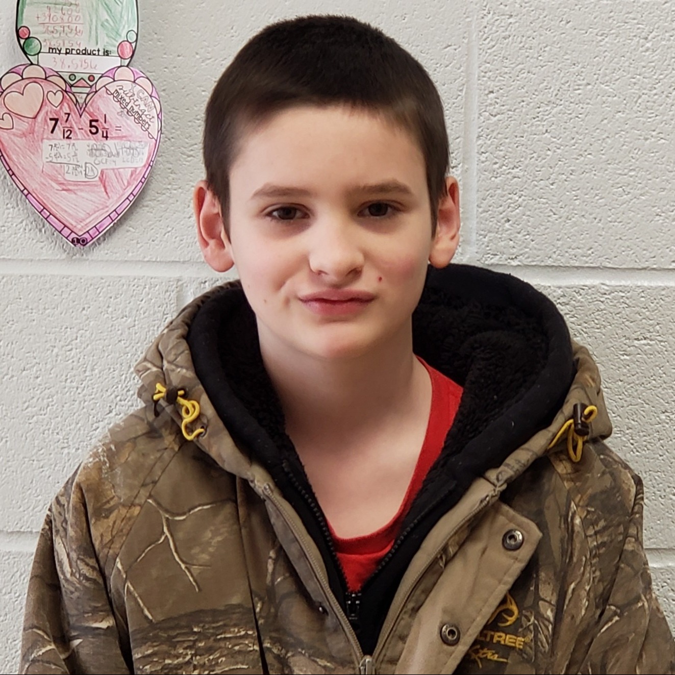 boy with dark hair wearing a red t-shirt under a zipped-up camouflage coat with hood