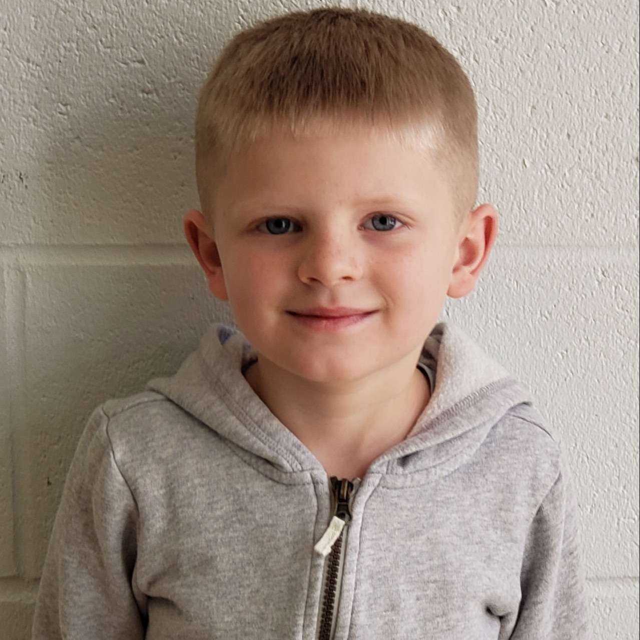 smiling young boy with blonde hair wearing a gray zipped up jacket