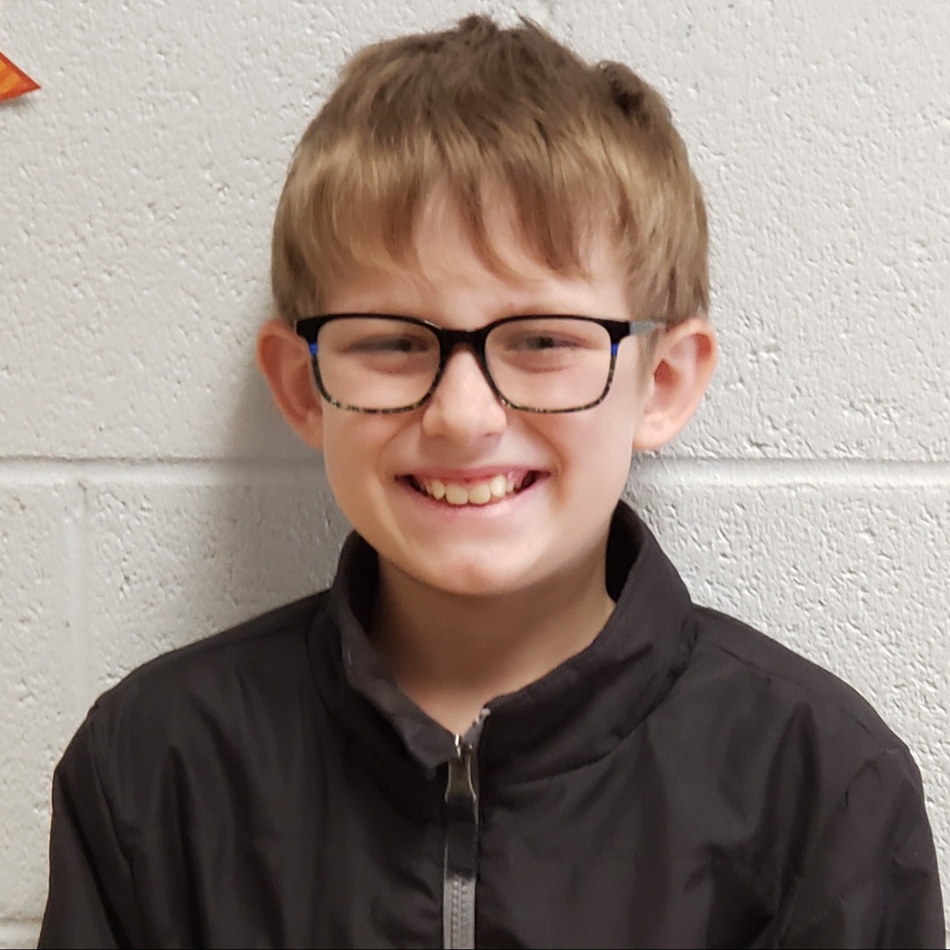 smiling boy with dark brown hair and glasses wearing a black  zipped up jacket