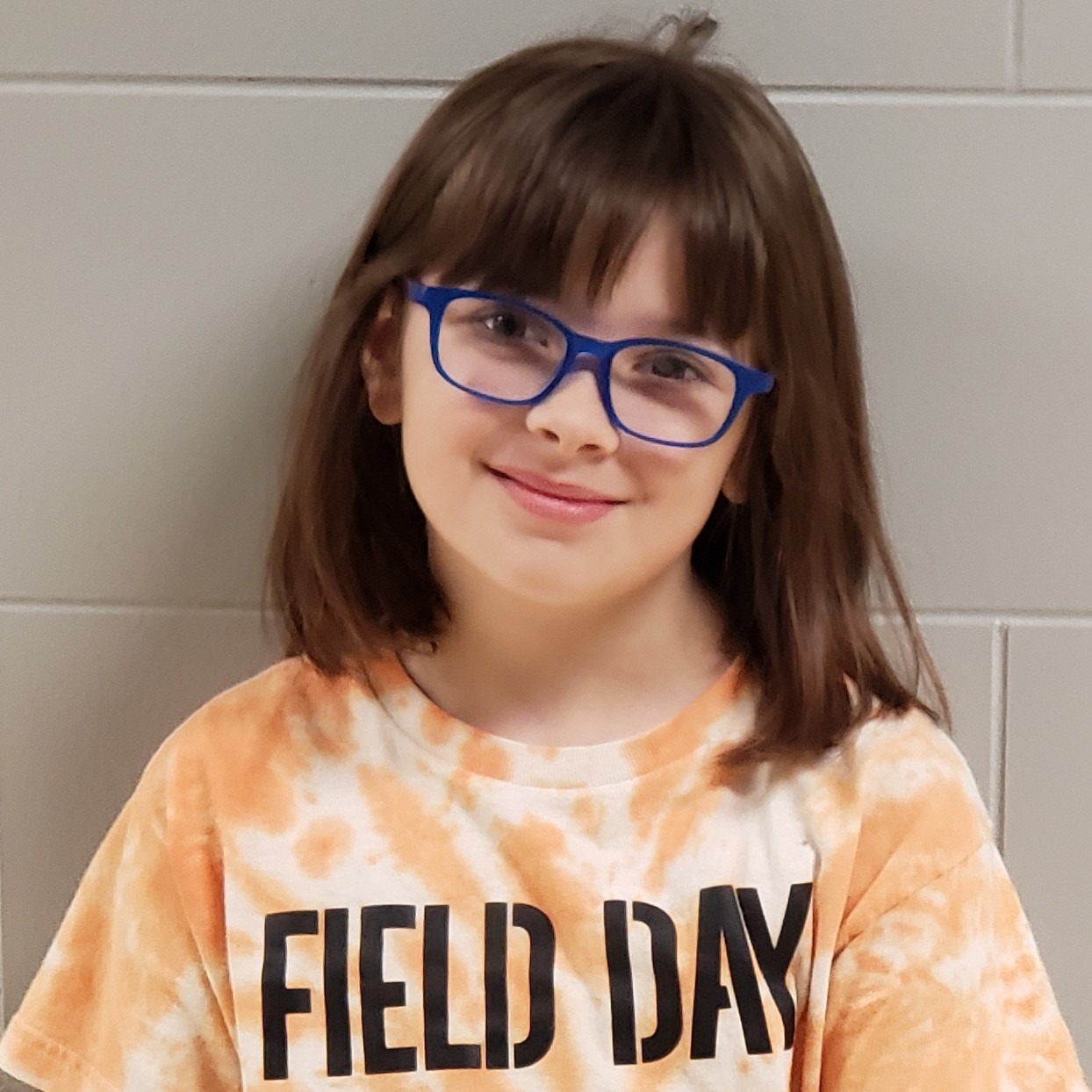 young smiling girl with dark hair and blue glasses wearing an orange tie-dyed FIELD DAY t-shirt