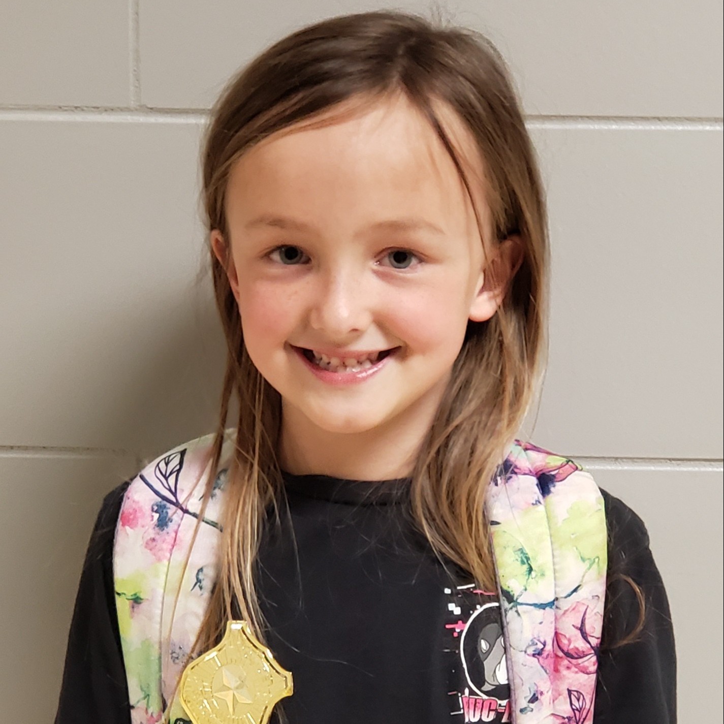 young girl with dark bonde hair wearing a black t-shirt, flowered backpack, and gold badge