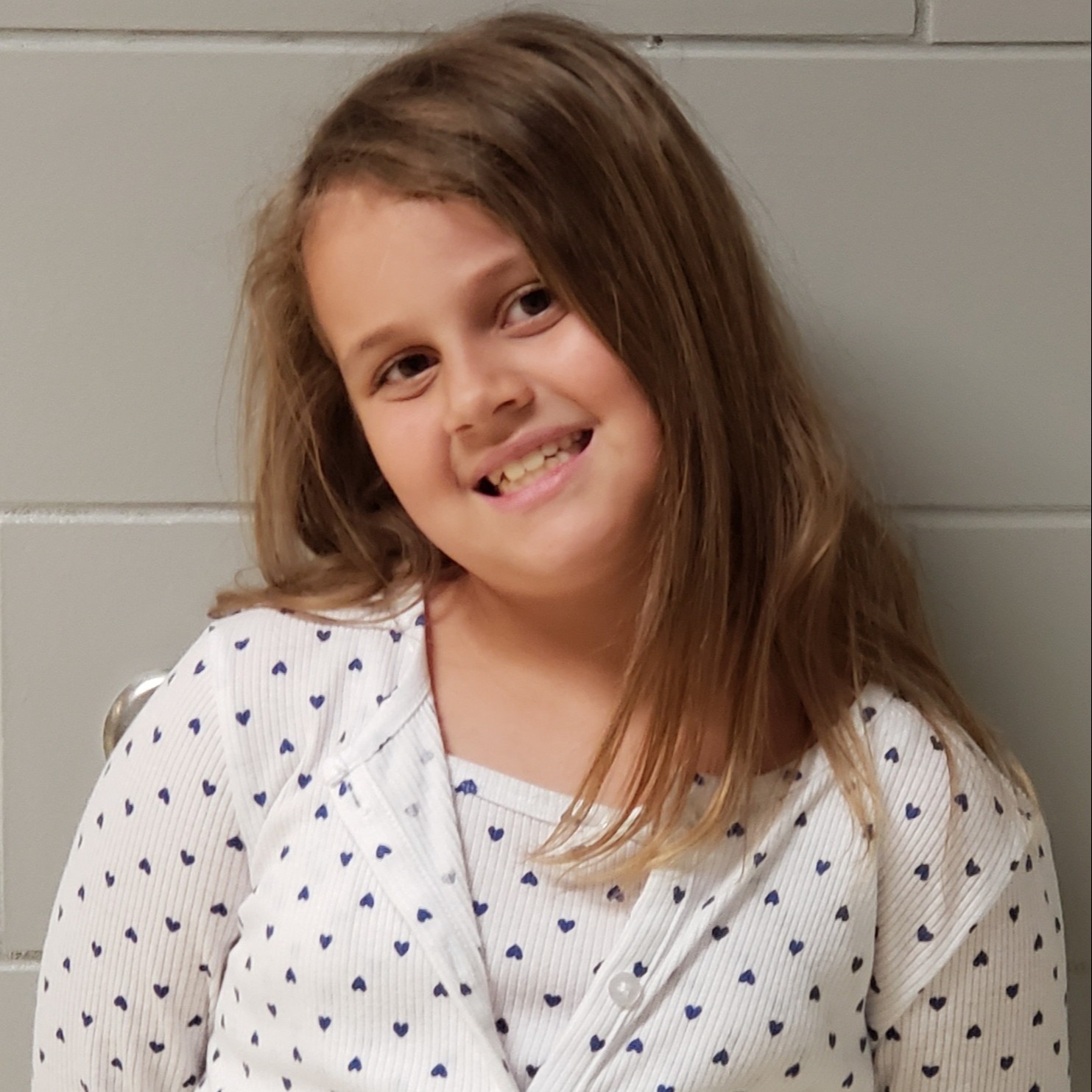 smiling young girl with dark blonde hair wearing a white shirt and cardigan with little blue hearts all over them