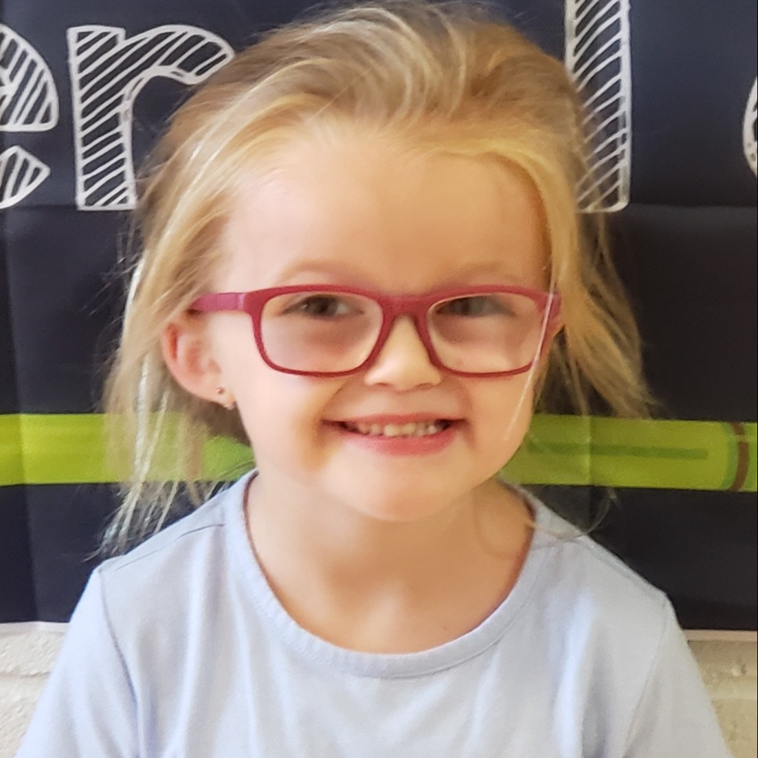 smiling little girl with blonde hair pulled up wearing pink glasses and a light blue t-shirt