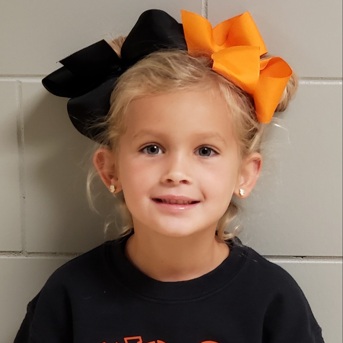 smiling little girl with blonde hair pulled up in orange and black bows wearing a black sweatshirt