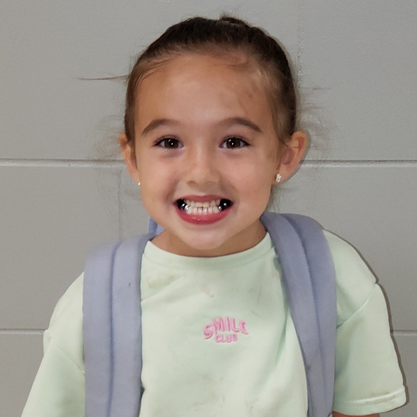 smiling little girl with brown hair pulled back wearing a light green sweatshirt and gray backpack