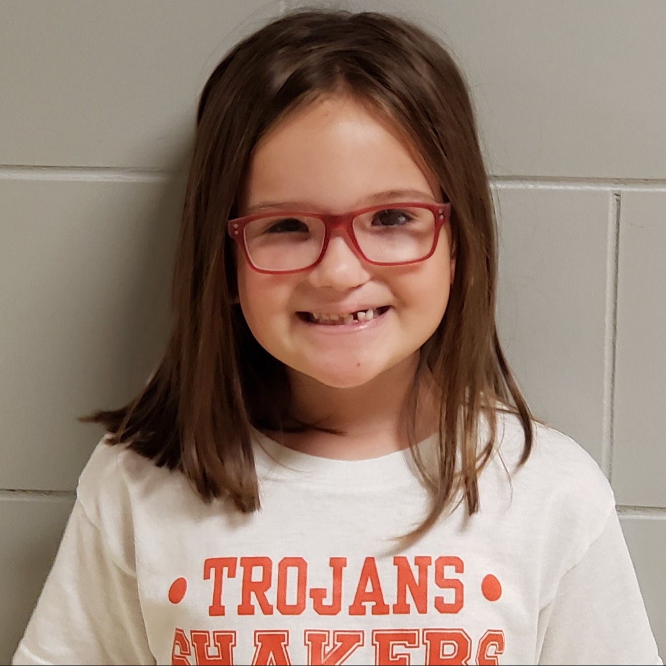 young girl with brown hair and red glasses wearing a white Trojans Shakers t-shirt