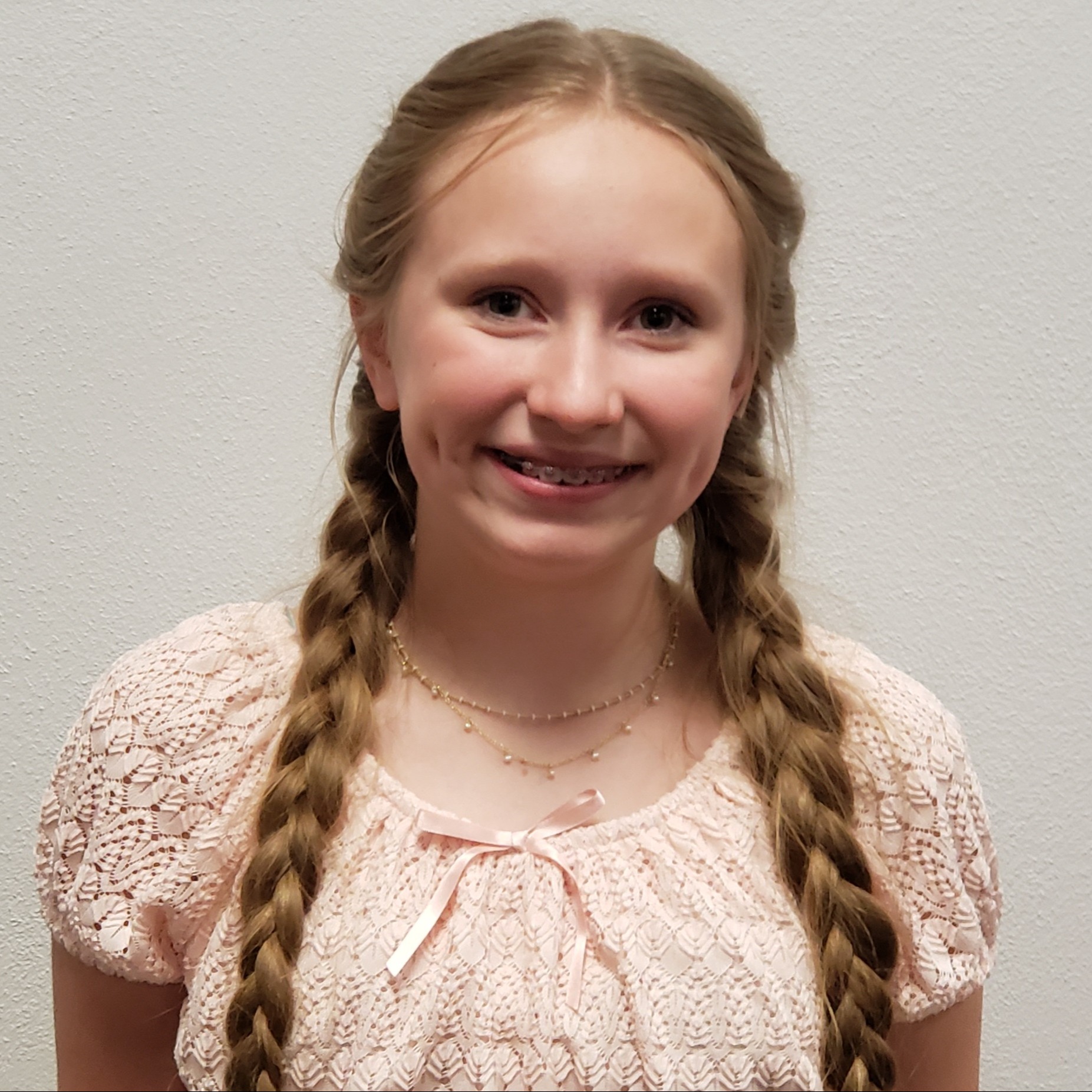 smiling girl with braces and blonde hair in braids wearing a pink lacy blouse and gold necklace