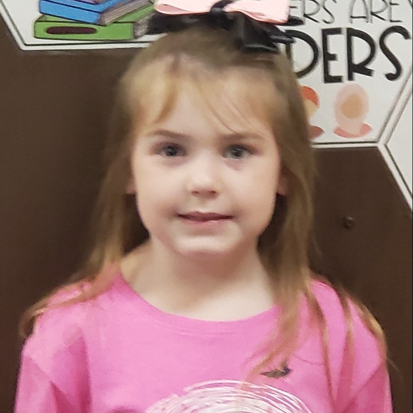 young girl with light brown hair in a black bow wearing a pink t-shirt