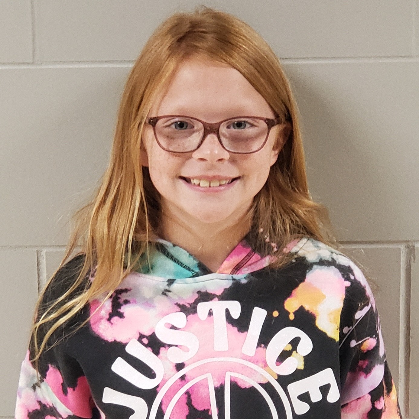 smiling girl with red hair, freckles, and brown-rimmed glasses wearing a tye-dyed Justice hoodie