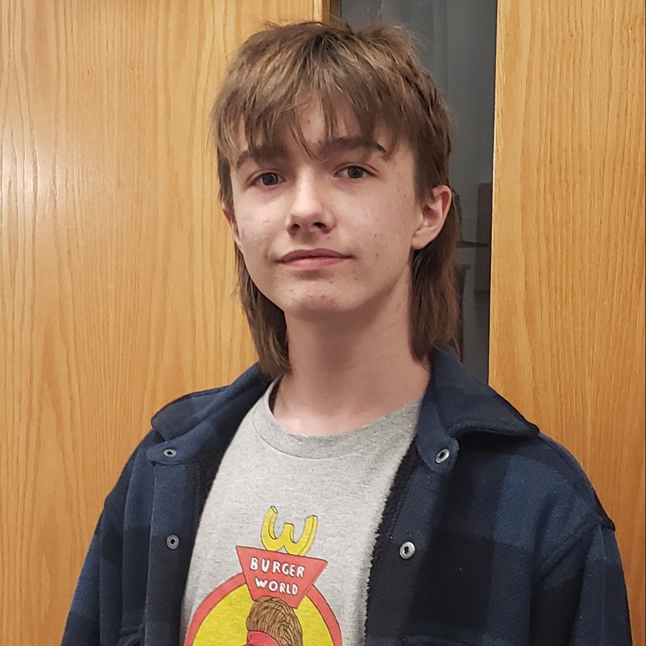 boy with a light brown mullet wearing a gray Burger World t-shirt under a navy blue plaid shirt