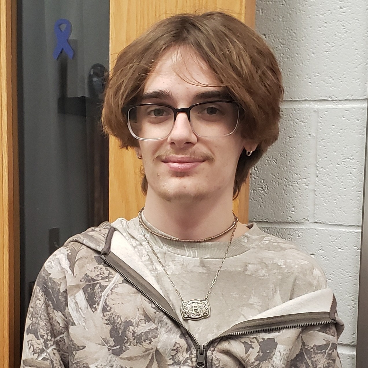 young man with reddish brown hair and mustache wearing glasses and a camo t-shirt under a camo jacket
