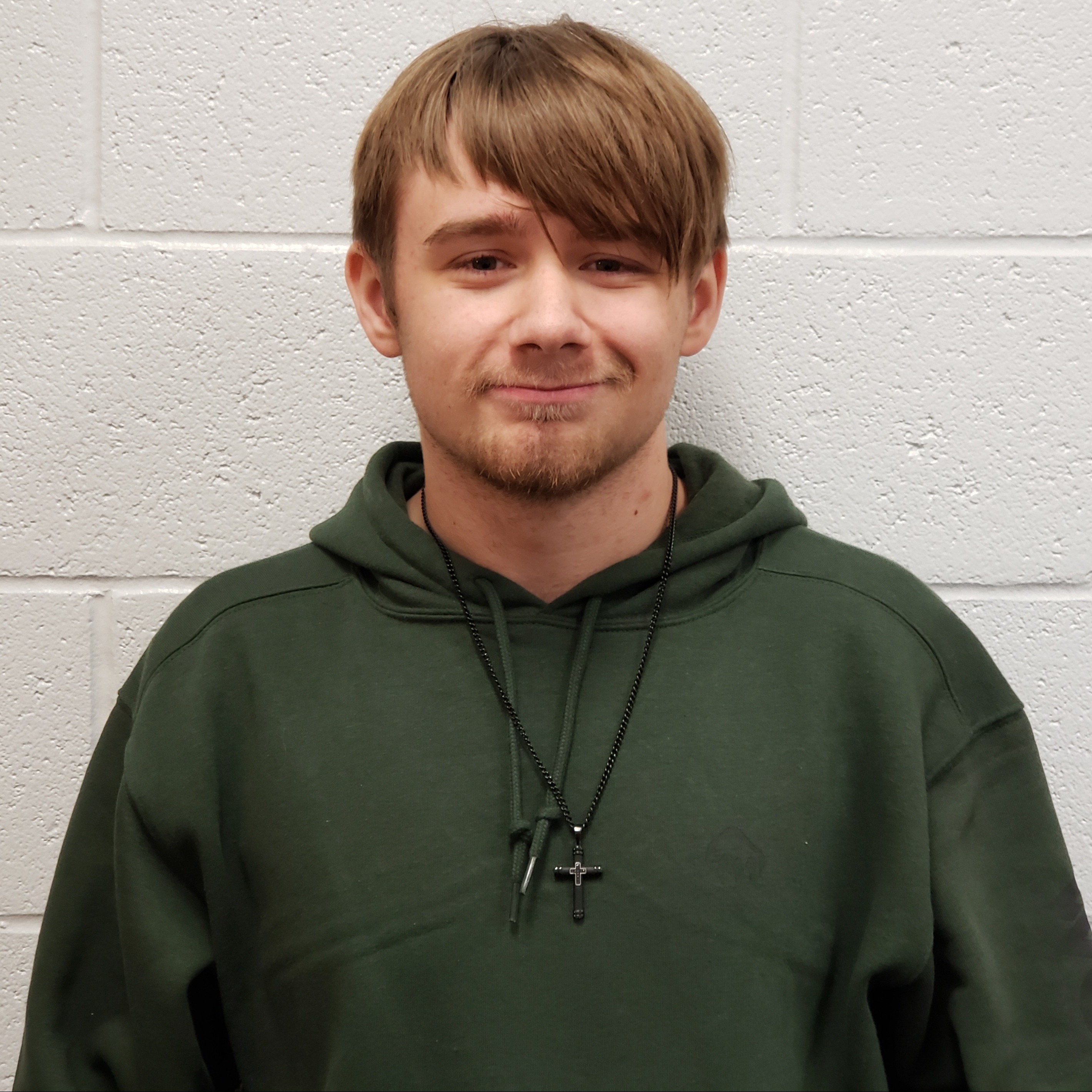 smiling young man with light brown hair and beard wearing a dark green hoodie with a cross necklace