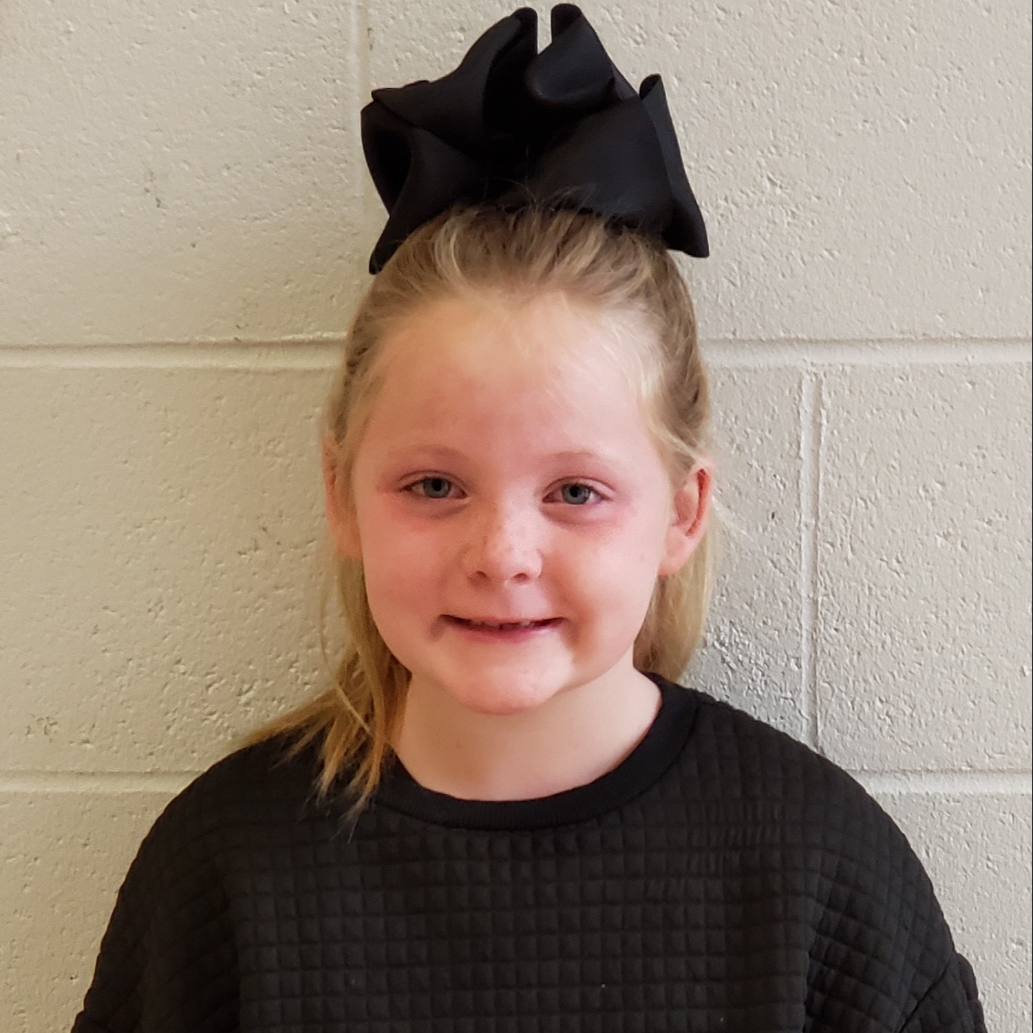 smiling young girl with blonde hair pulled up in a black bow wearing a black shirt