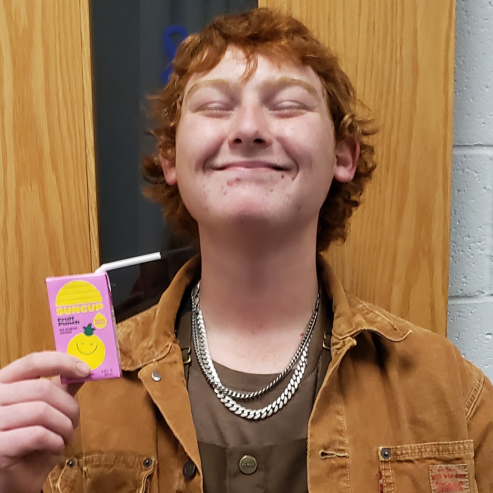 smiling young man with red hair and gold necklaces wearing a brown canvas jacket over a brown t-shirt and holding up a pink juicebox