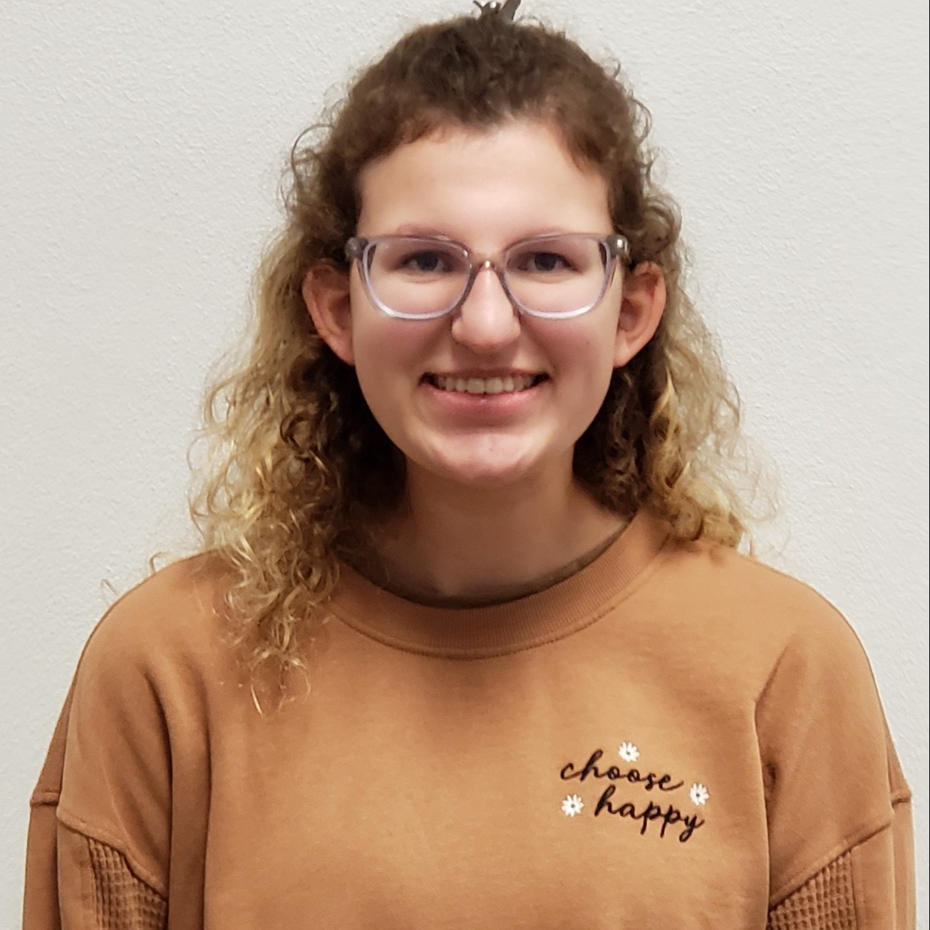 smiling young lady with curly blonde hair and glasses wearing a light brown "choose happy" sweatshirt