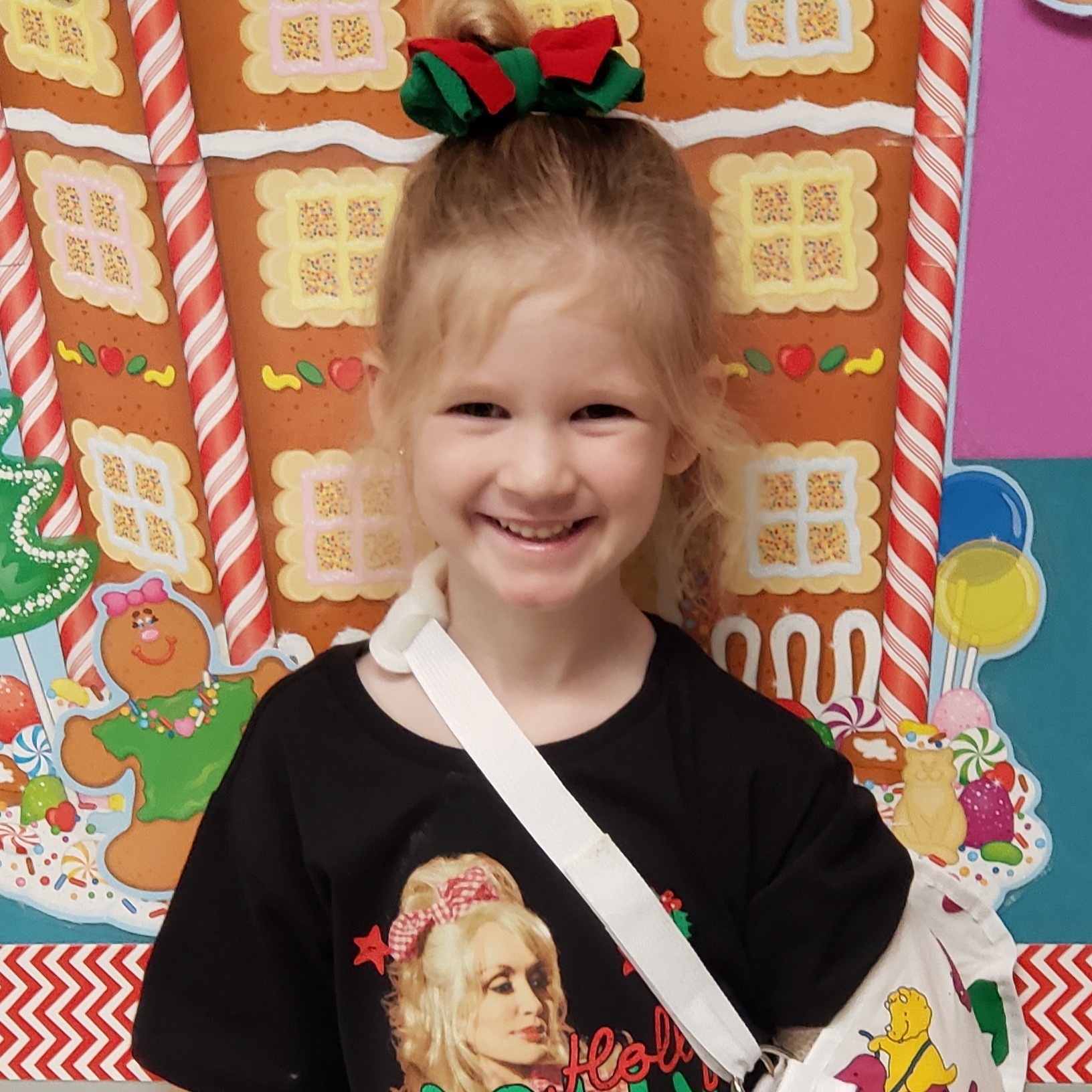 smiling little girl with blonde hair pulled up in a red & green bow wearing a black Dolly  t-shirt and a white sling with  yellow, pink, & green dinosaurs on it