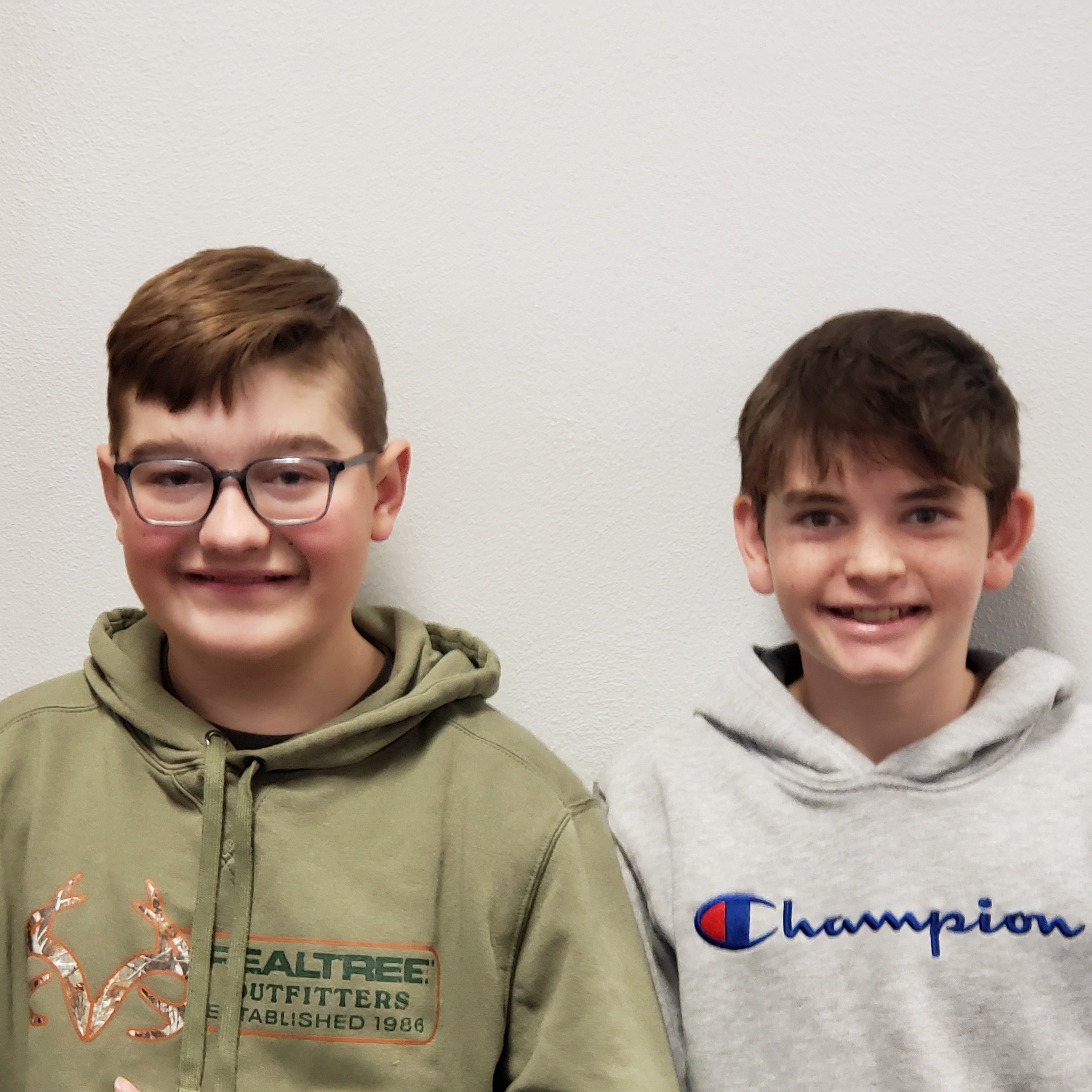 2 smiling boys, one with light brown hair and glasses wearing a light green Realtree hoodie, the other with dark hair wearing a gray Champion hoodie