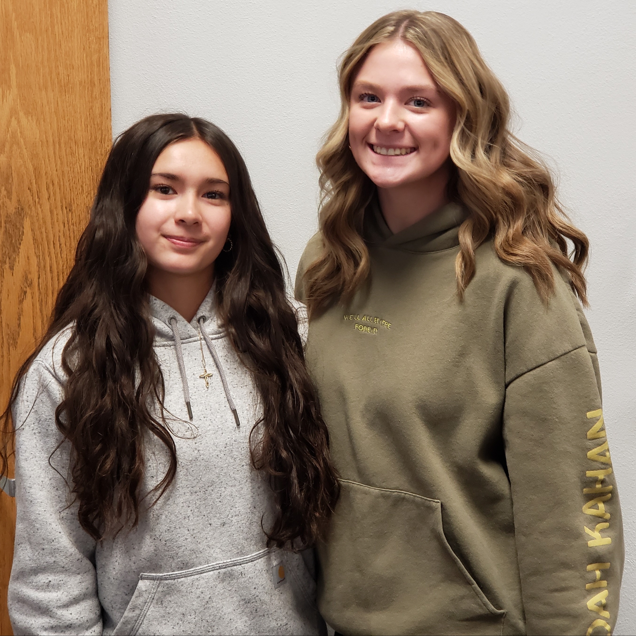 2 smiling young ladies, one with long dark hair wearing a gray hoodie and gold cross necklace, the other with blonde hair wearing a tan hoodie