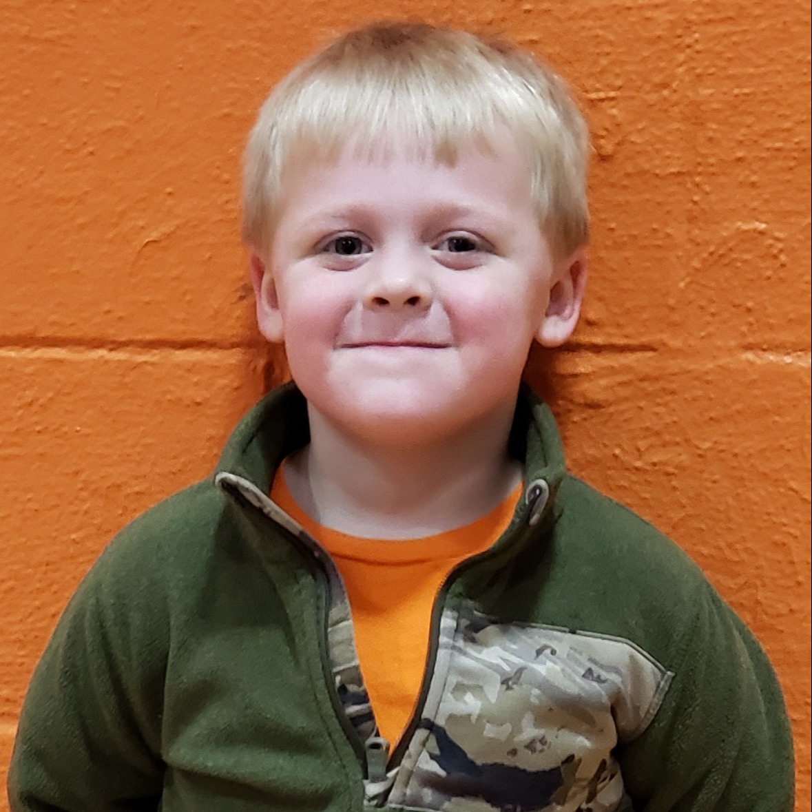 smiling little boy with light blonde hair wearing an orange t-shirt under a green 3/4 zip sweatshirt