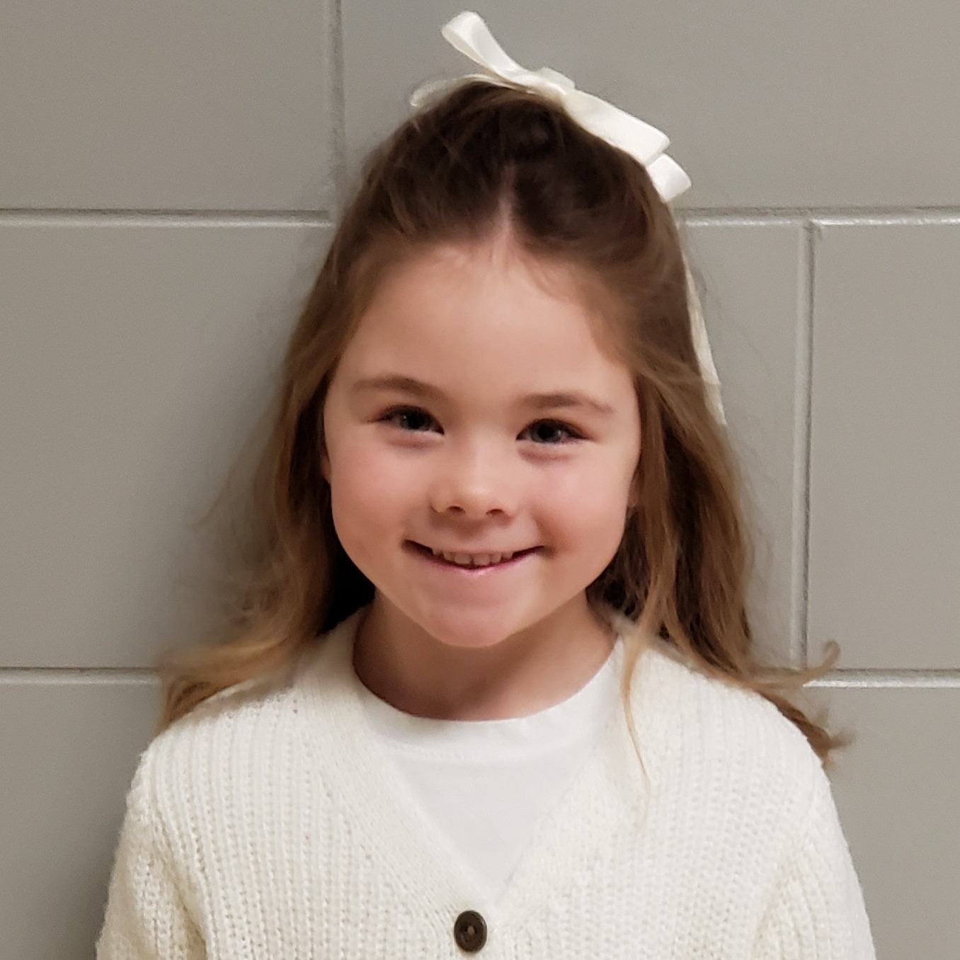 smiling girl with dark blonde hair pulled half up in a white bow wearing a white cardigan over a white shirt