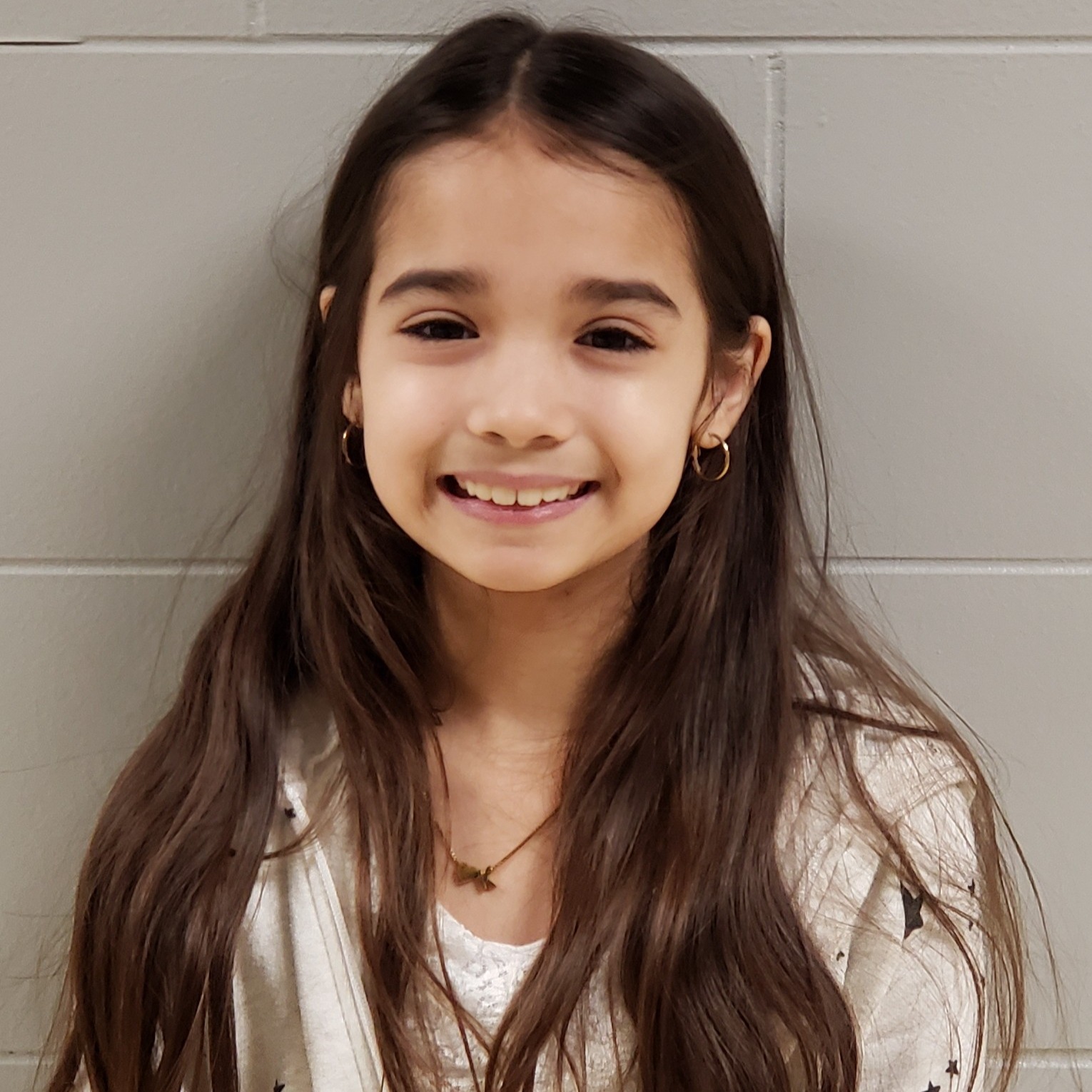 smiling girl with long brown hair wearing hoop earrings and a white zip-up jacket with blue stars over a white lacy shirt