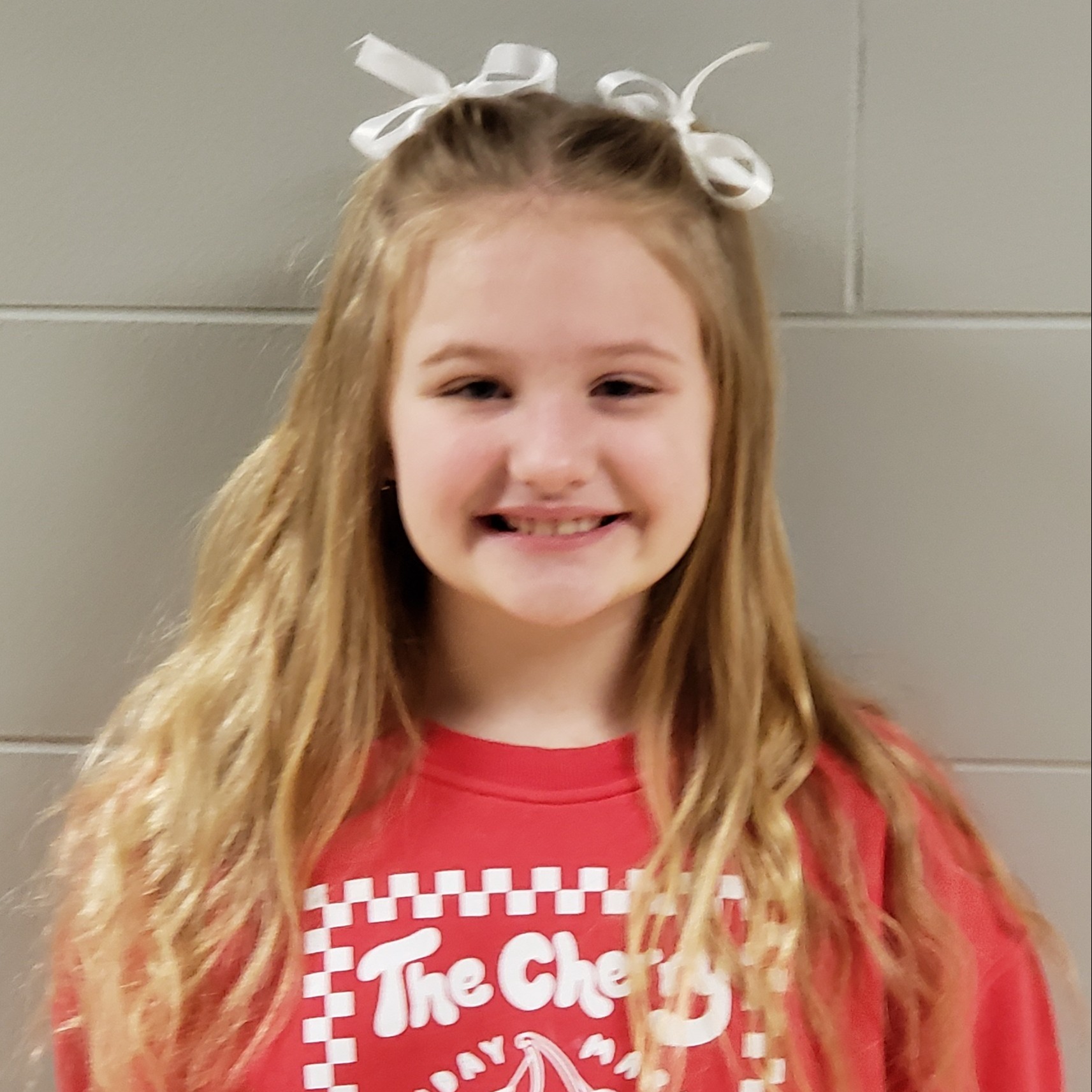 smiling girl with long blonde hair pulled up in white bows wearing a red sweatshirt