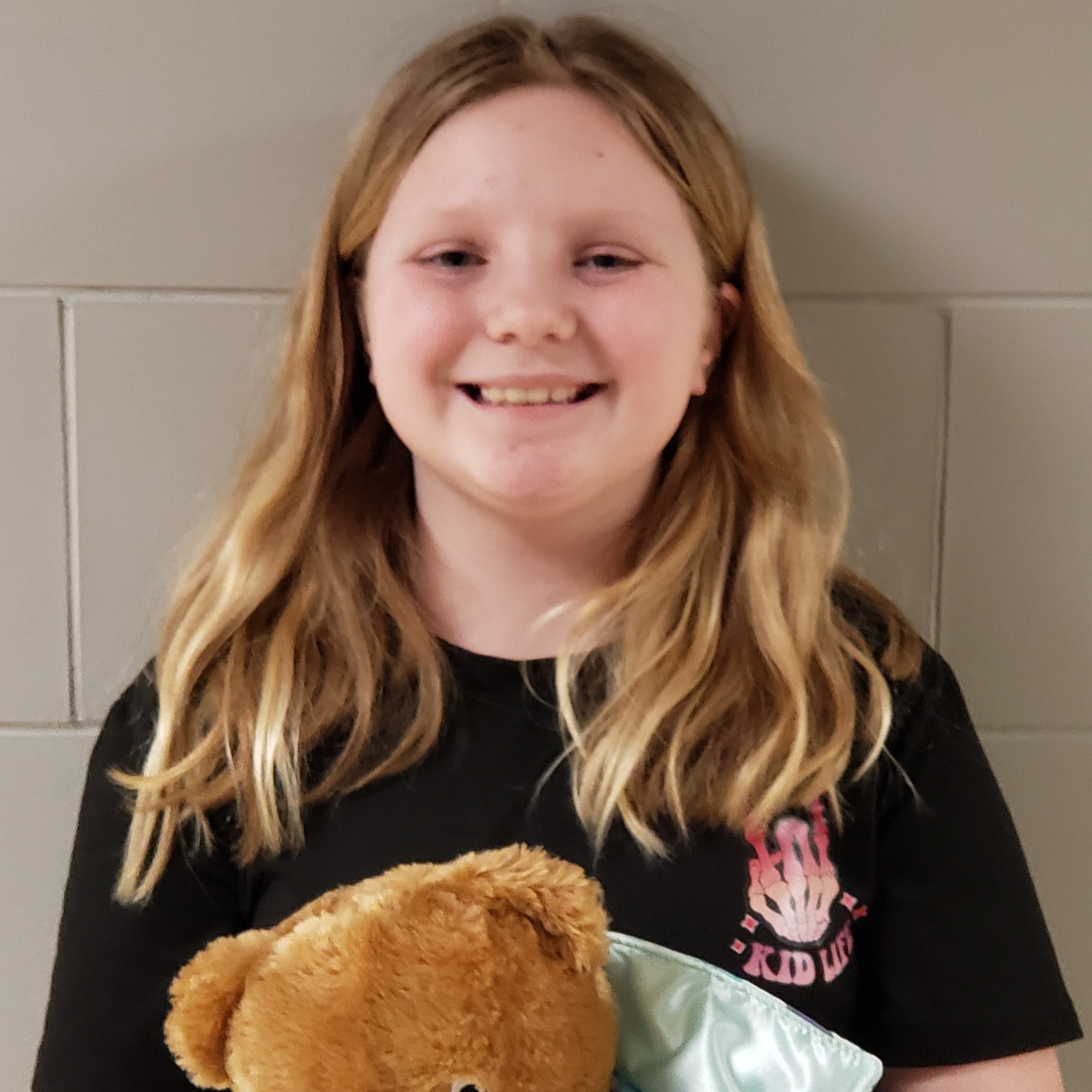 smiling girl with long blonde hair wearing a black t-shirt and holding a teddy bear