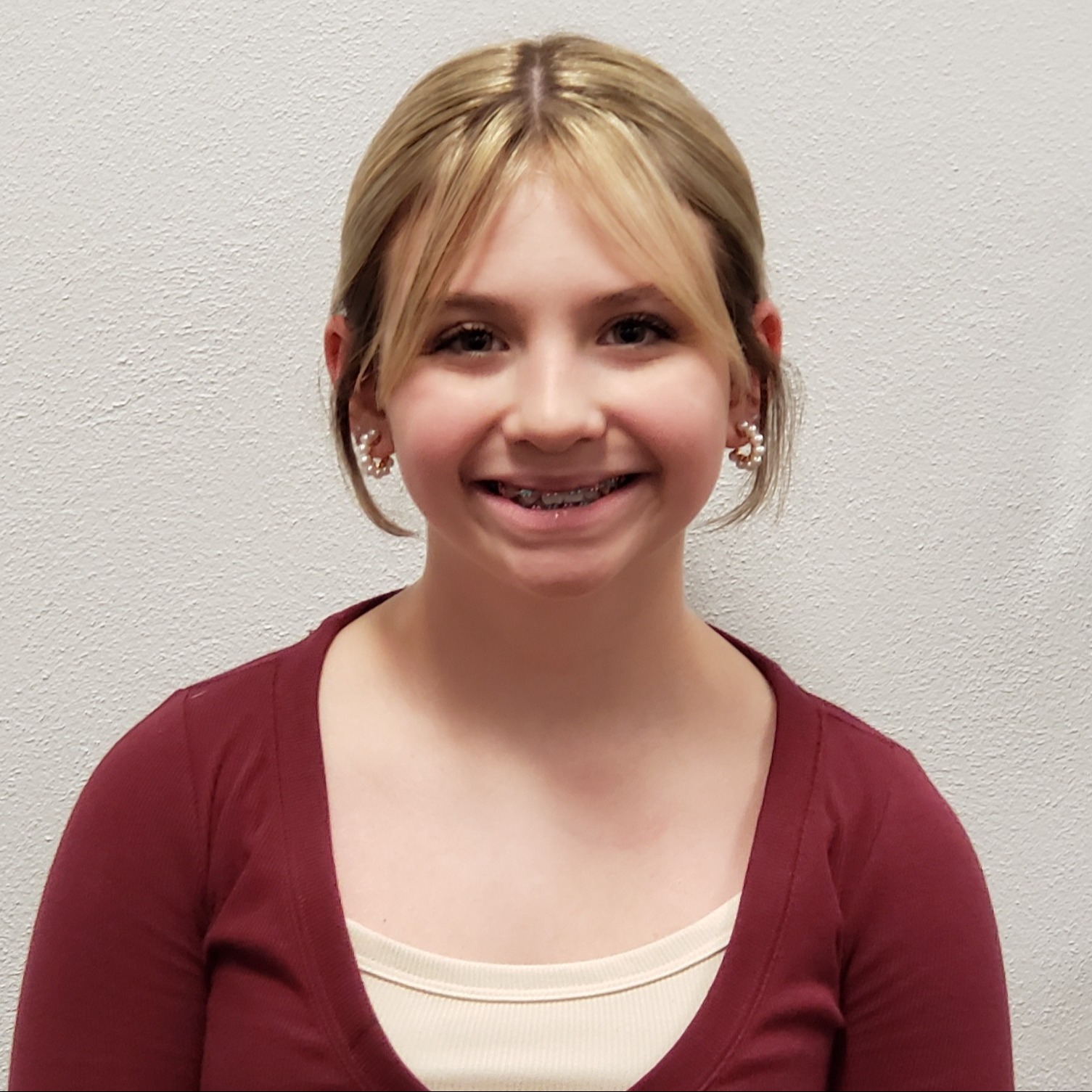 smiling girl with blonde hair pulled back, pearl hoop earrings, and braces wearing a maroon shirt over a tan undershirt