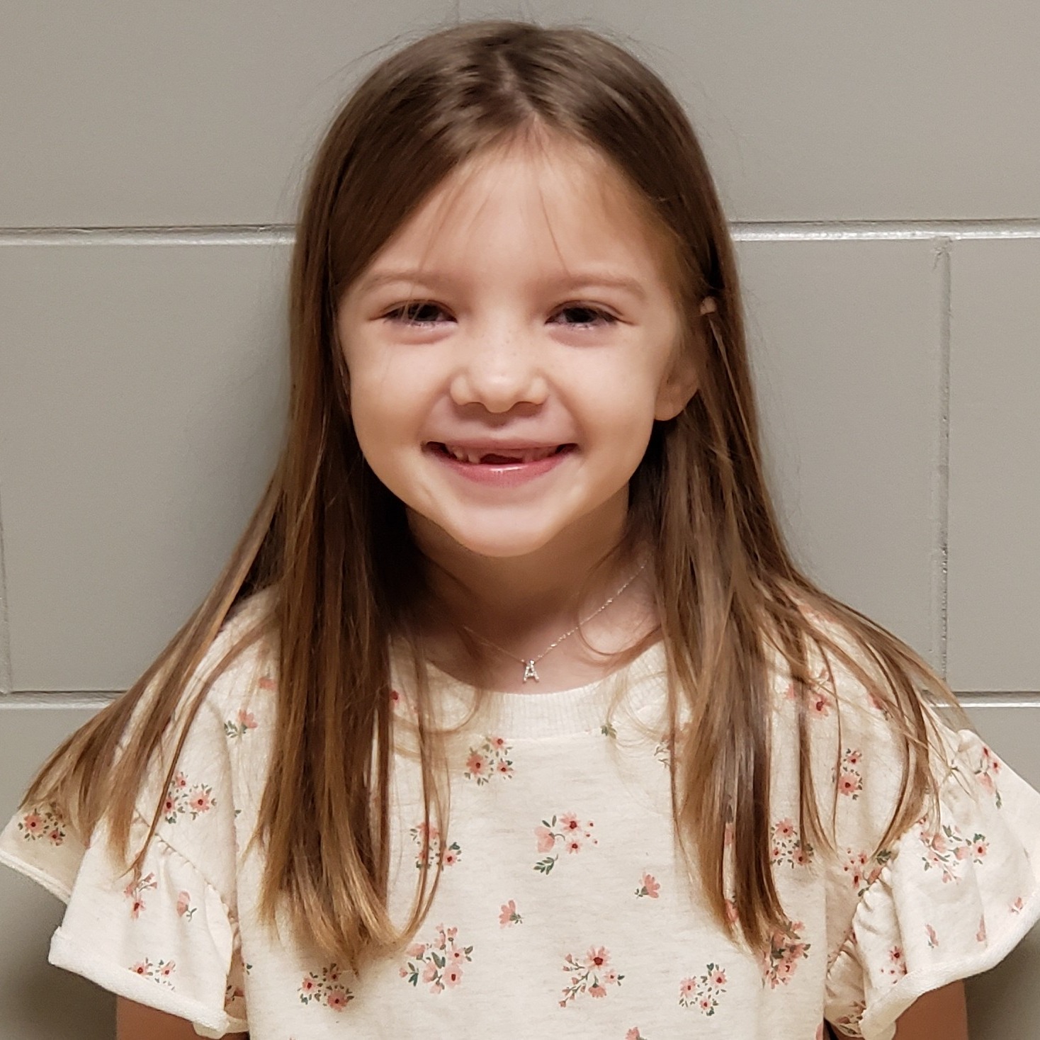 smiling girl with long dark blonde hair, missing her front teeth, and wearing an "A" necklace and cream shirt with ruffled sleeves and flowers on it