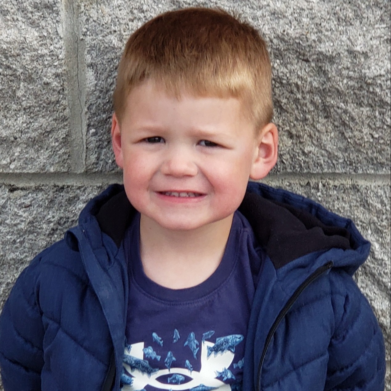 smiling little boy with blonde hair wearing a blue t-shirt under a blue coat